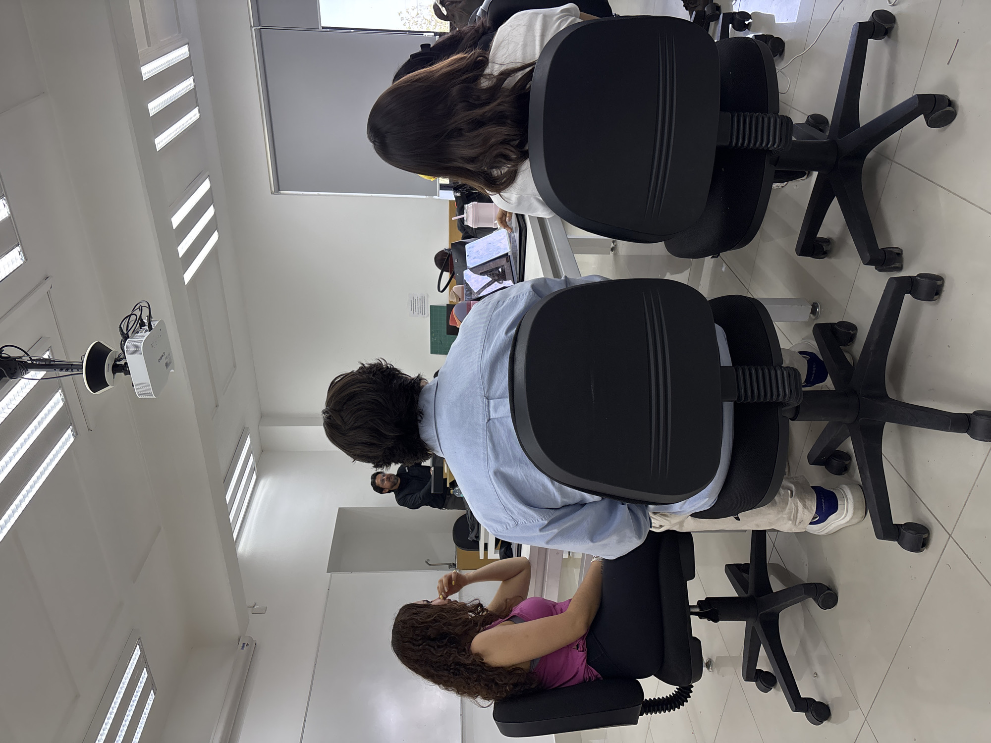 The image depicts an indoor classroom setting in San Andrés Cholula, Mexico. Three individuals are visible, seated on black office chairs around tables. The foreground subject, a woman with curly brown hair, wears a pink top and black pants and has her hand near her mouth. Behind her, a person in a light blue shirt is seated at a desk. Further away and in front, a person with long brown hair wearing a white shirt is also sitting at a desk, with a cup with a straw on the table. Tables with various items are partially visible. A projector hangs from the white ceiling, which is illuminated by linear lights. The floor is tiled. More people can be seen further back.