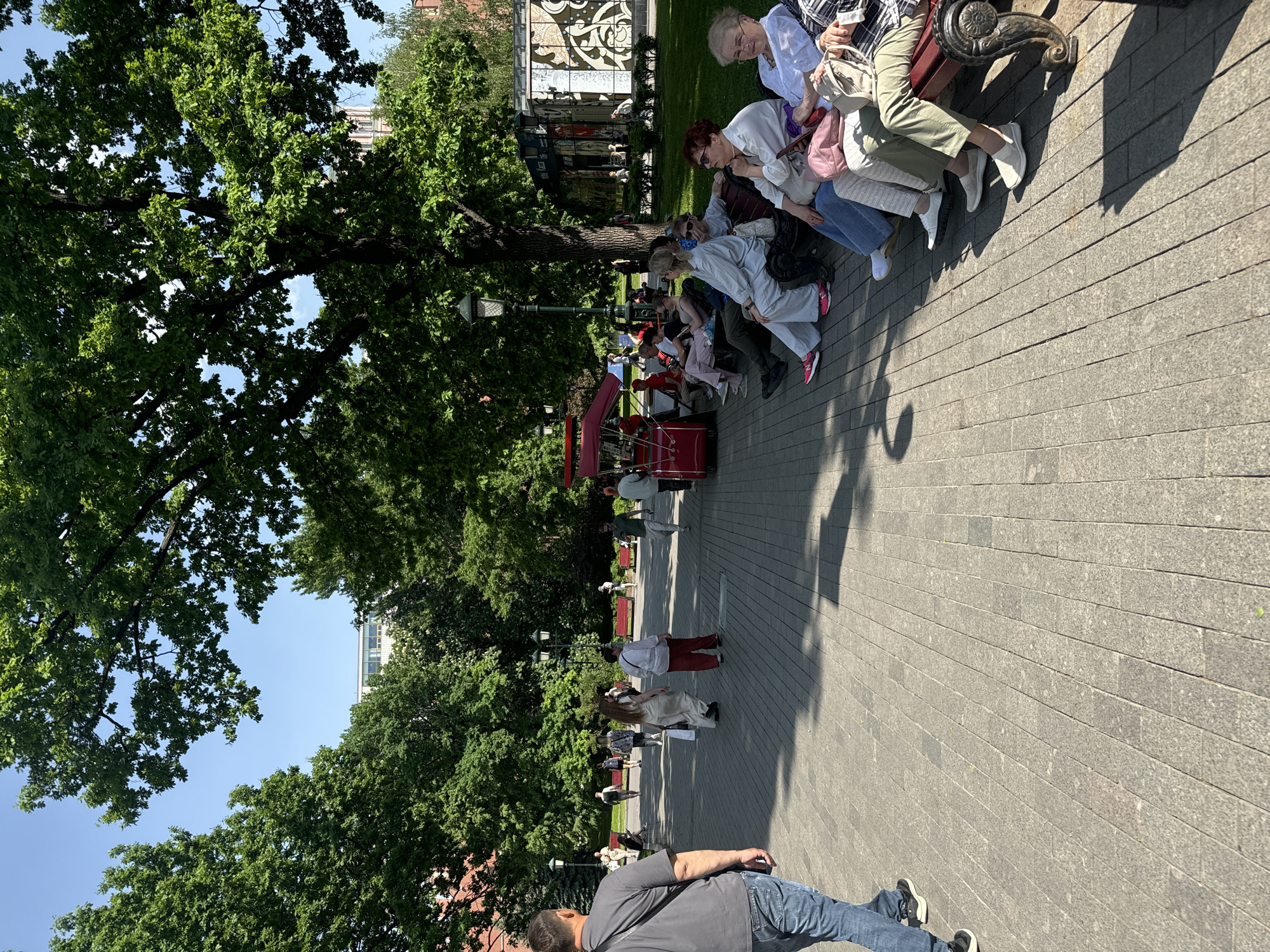 The image shows a park in Moscow, Russia. The ground is paved with gray rectangular stones. A large tree with dense green foliage dominates the left side of the frame, casting shadows on the ground. To the right of the tree, a line of people is seated on a bench. The people vary in age and attire. Further down the path, there are other people standing and walking. A red cart with a canopy is visible in the distance. A man wearing a gray shirt and jeans is walking towards the camera in the foreground. In the background, there is a building with a decorative facade. The sky is blue and mostly clear.