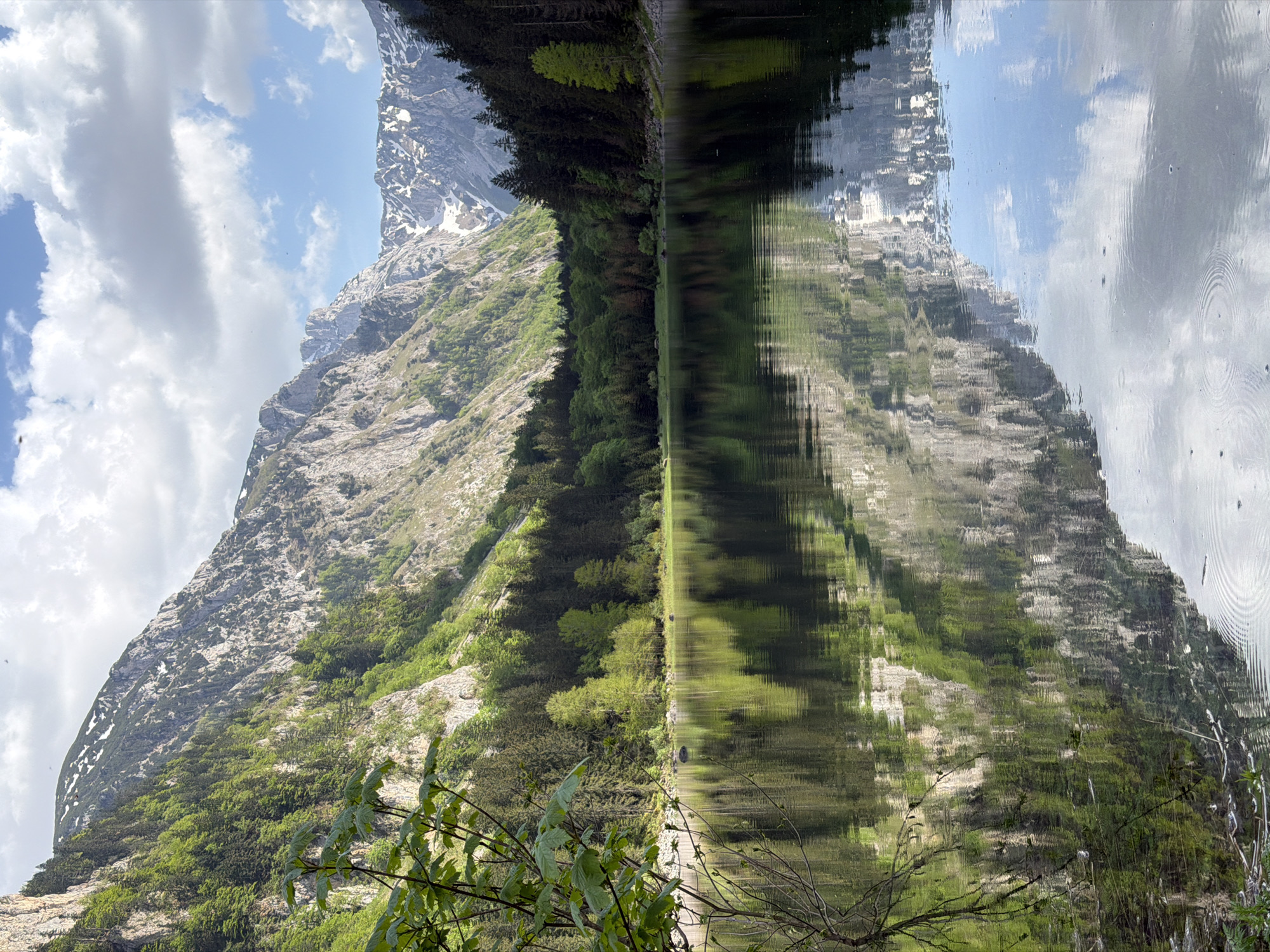 The image depicts a mountain and its reflection in water, likely a lake. The mountain is rocky with patches of snow near the top and is covered with greenery at its base. A line of trees separates the mountain from the body of water. The water is calm enough to create a clear reflection of the mountain, sky, and clouds. The presence of mountain scenery and water suggests a natural setting, which might correspond to the area near Pluzine, Montenegro. The photograph seems to emphasize the natural beauty of the mountain landscape and its reflection.
