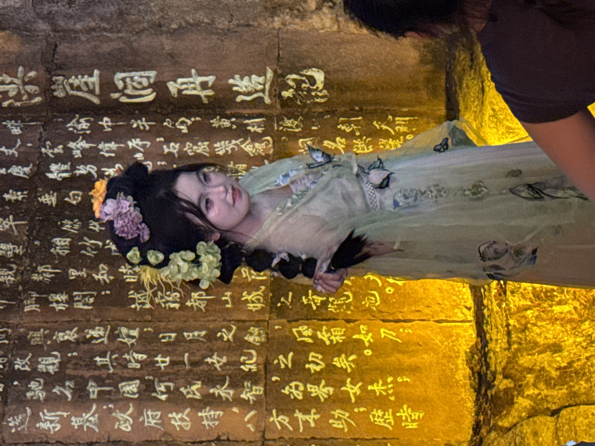 A young woman with elaborate floral hair ornaments and makeup stands facing right in front of a stone wall inscribed with golden Chinese characters. She wears a light green, translucent robe with floral motifs. The stone wall is illuminated by a warm, yellow light, highlighting the intricate calligraphy. The setting appears to be outdoors in a rocky, uneven terrain, possibly in the Yuzhong District of China, known for its historical sites and mountainous landscape. The woman's pose suggests she is a performer or participant in a cultural event.