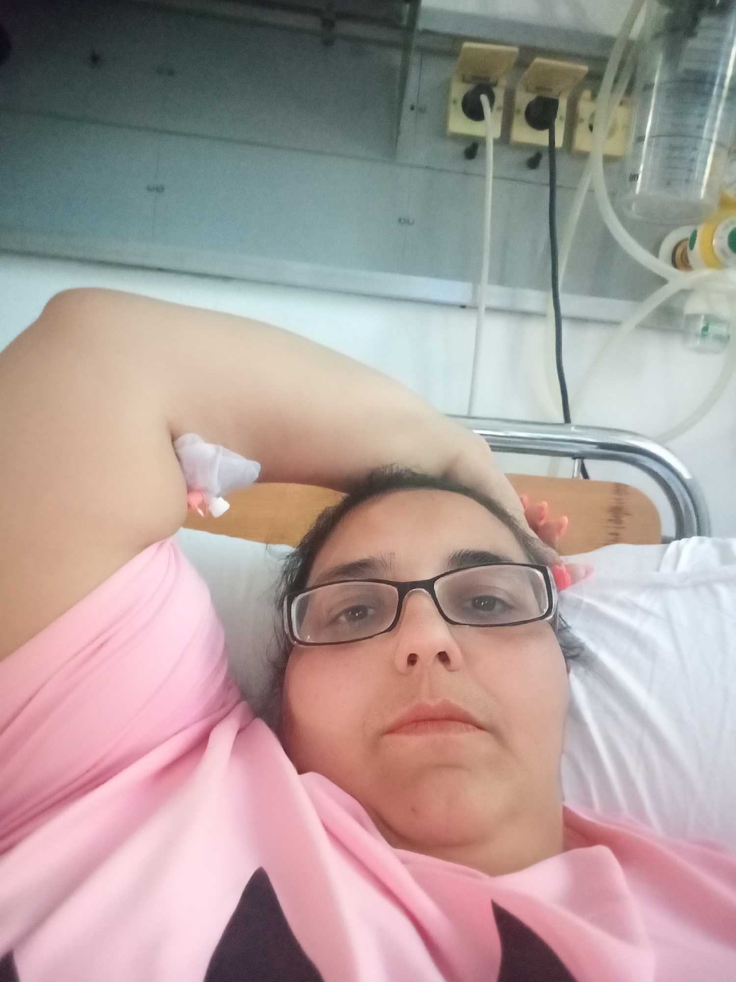 The image shows a selfie taken by a person in a hospital bed. The person is wearing glasses and a pink top. The background appears to be a hospital room with medical equipment visible, including an IV drip and what look like electrical outlets for medical devices. The image does not contain enough visual information to determine the specific location in Pleven, Bulgaria.
