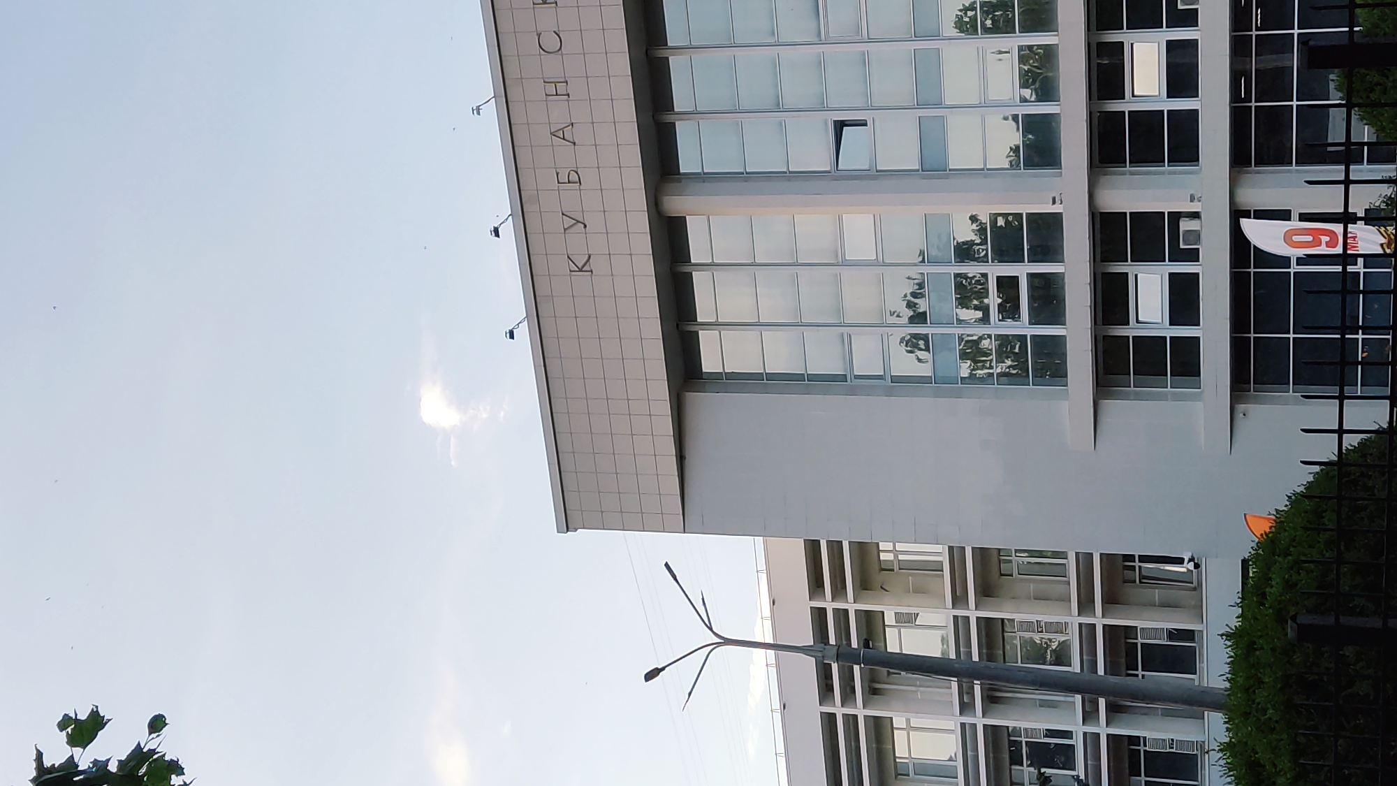This is a daytime, exterior shot of a modern building in Krasnodar, Russia. The building features a grid-like facade with numerous windows, many of which reflect the sky and surrounding trees. The name "КУБАНЬ" is visible in large letters across the top of the building. A black metal fence runs along the bottom of the frame, partially obscuring a well-maintained hedge. A street lamp with multiple arms is visible in the foreground, and a few small birds are scattered in the sky. A bright orange banner with the number "6" is visible on the right side of the building, near the fence. The sky is light blue with a few wispy clouds, suggesting fair weather. The overall impression is of an urban, institutional setting on a clear day.
