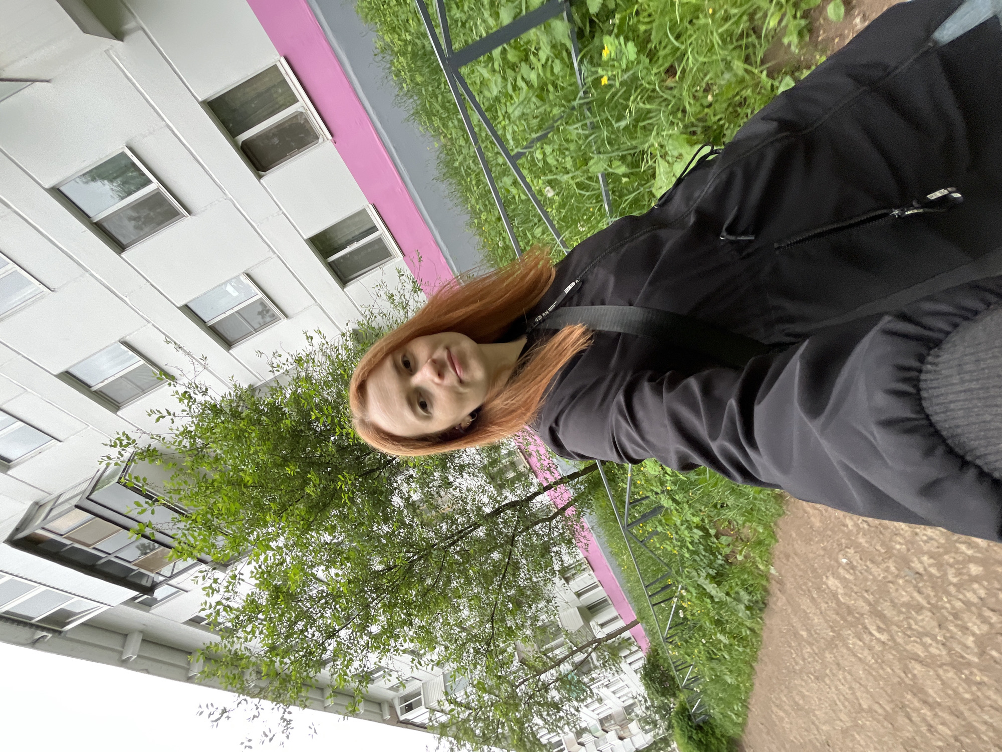A fair-skinned woman with long red hair wearing a black jacket and gray undershirt is standing outside a multi-story, residential building with white and pink facade and multiple windows. The building appears to be located in the city of Chaikovsky, Russia. A metal fence runs parallel to a dirt path, bordering an area of grass and weeds. A tree is in the foreground, partially obscuring the view of the building.