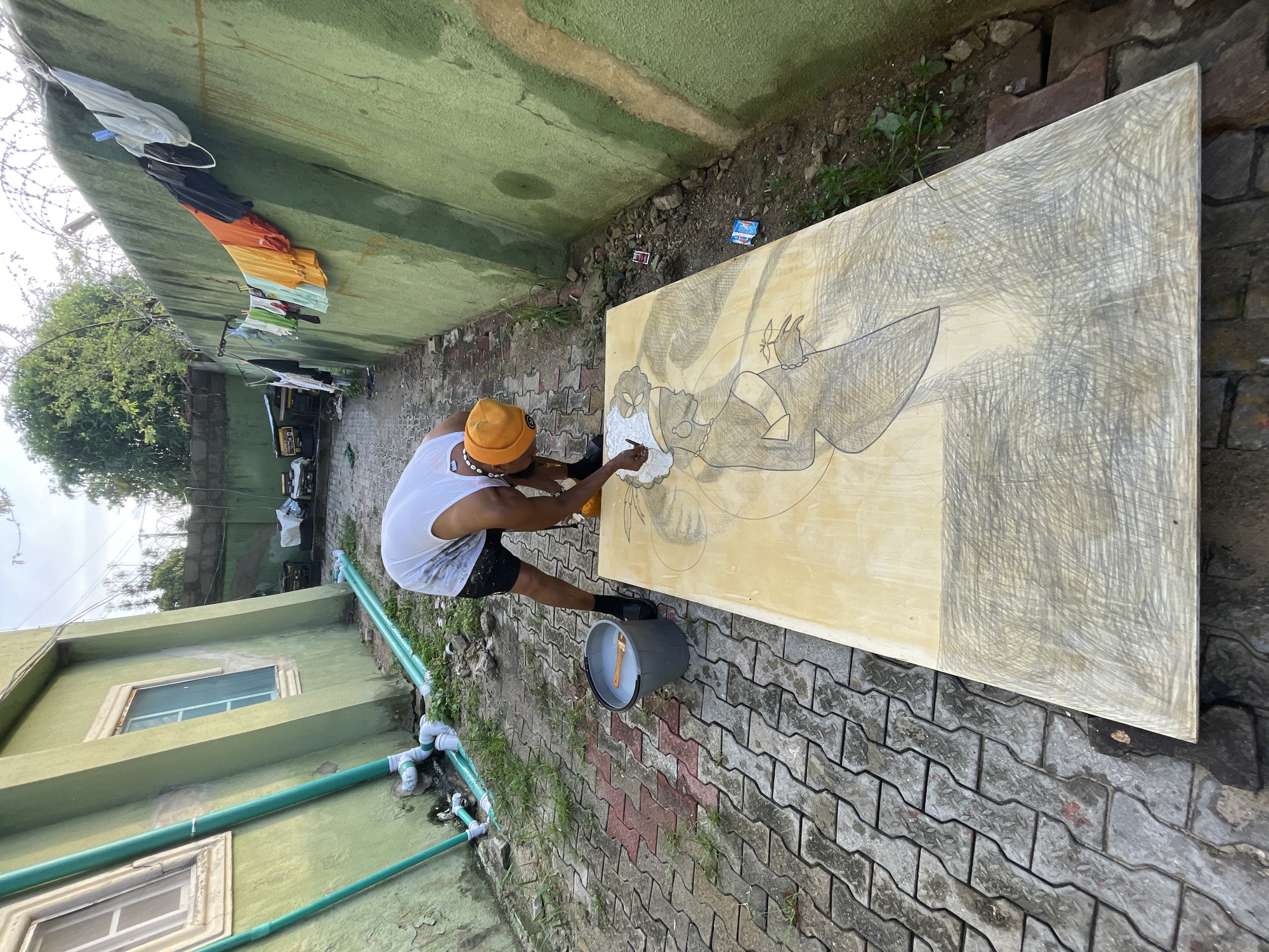 A man stands in an outdoor area painting on a large canvas. He wears a yellow beanie, a white tank top, and dark shorts. The canvas is positioned on a brick-paved surface, displaying a sketch of a figure. To the man's right is a grey bucket containing a paintbrush. Clothes hang on a line above the canvas and a green building stands in the background. The outdoor area appears to be located in Ado, Nigeria. The area includes green drainage pipes, vegetation and an electrical generator can be seen in the background. A white packet and red and white box of matches lay on the ground behind the canvas.