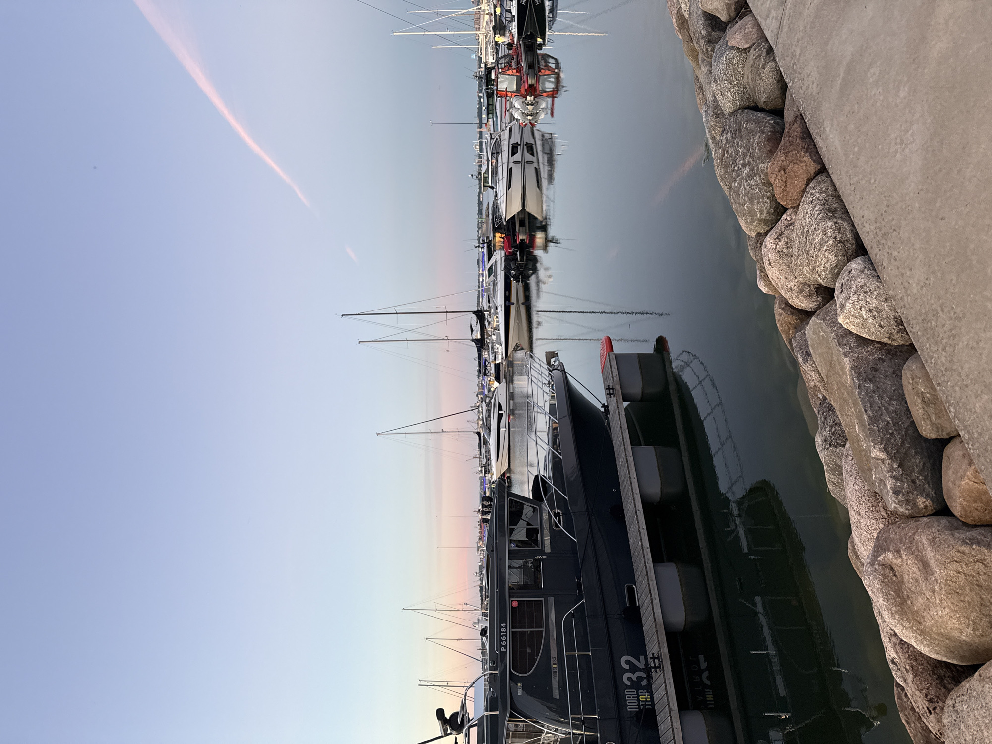 The image depicts a marina in Tallinn, Estonia, during twilight. A dark-colored motorboat, identified as "NORD STAR 32", is docked prominently in the foreground, its reflection visible in the calm water. Beyond this vessel, numerous sailboats and other motorboats are moored in the harbor. The sky is a soft gradient of blue, transitioning to pink hues near the horizon, with a faint contrail visible. The shoreline is reinforced with large, rough-hewn rocks and a concrete embankment. The overall scene conveys a sense of tranquility and evening serenity in a maritime setting.