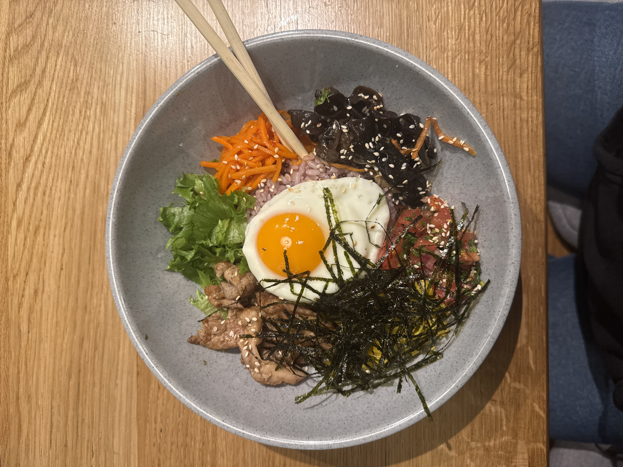 A close-up, overhead shot shows a grey bowl filled with a Bibimbap-like dish, potentially in Saint Petersburg, Russia (based on context clues not visible in the image). The dish includes purple rice, shredded carrots with sesame seeds, dark wood ear mushrooms, lettuce, seasoned meat (possibly pork), seaweed strips, an egg fried sunny-side up, and a small amount of shredded yellow vegetables. The bowl rests on a wooden table, with chopsticks leaning on the side. A person wearing jeans is seated next to the table.