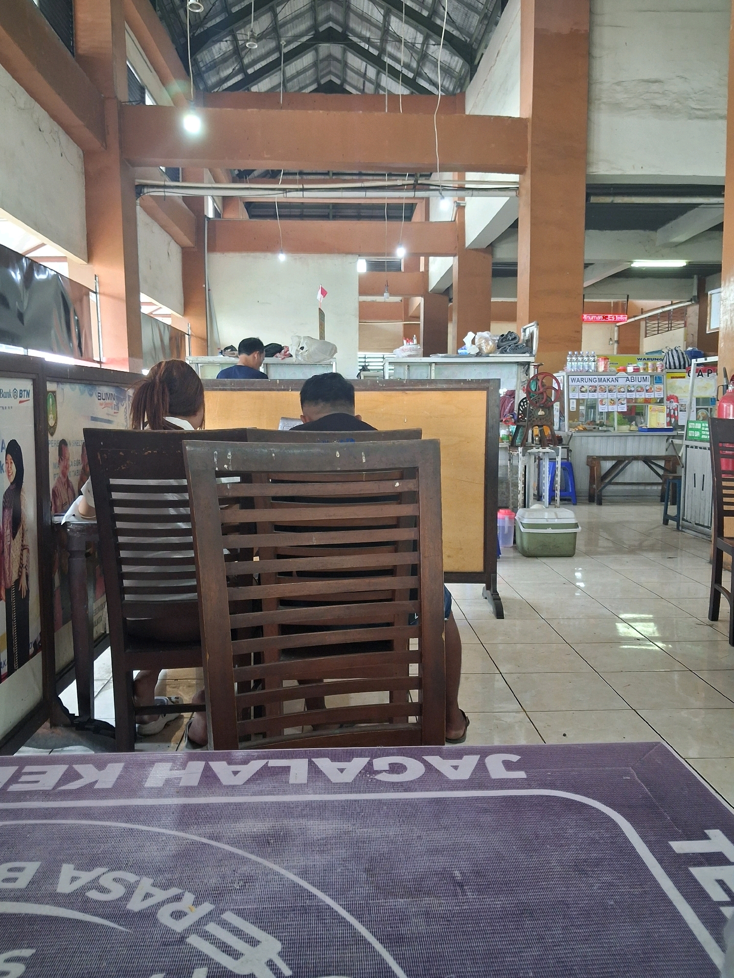 The image depicts an indoor food court or dining area, possibly in Surakarta, Indonesia. In the foreground, a purple mat with white lettering, partially obscured, is visible on a table. Behind it, two people are seated at wooden tables and chairs. One person, a woman with her hair in a ponytail, is facing away from the camera, leaning forward. The other, a man with short hair, is also facing away. Further back, a food stall with various items displayed is visible, including drinks and what appears to be a shaved ice machine. The overall environment is functional and somewhat dated, with concrete pillars and a metal roof structure. The lighting suggests it's daytime, with a mix of natural light and artificial illumination.