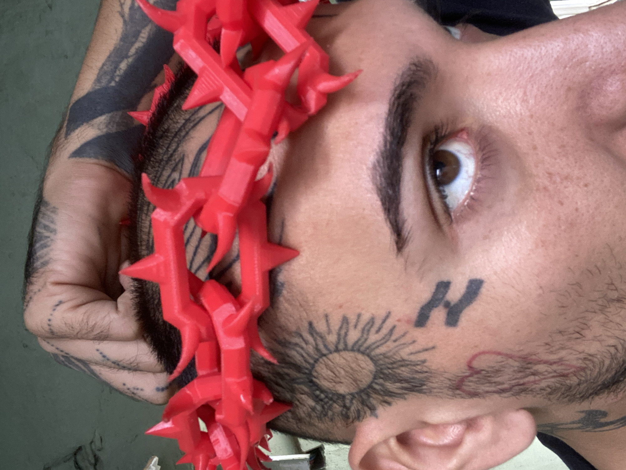 A close-up, tilted shot features a person wearing a red, plastic crown of thorns. The person has light brown skin, dark hair, and facial tattoos. These include a large sun-like design and the letter "N." The person's eyes are brown. Their arm, which has dark-colored tattoos, is visible, holding the crown in place. The background is blurry and indistinct, but it appears to be an indoor setting with pale green walls. The location is likely not visible in the image.