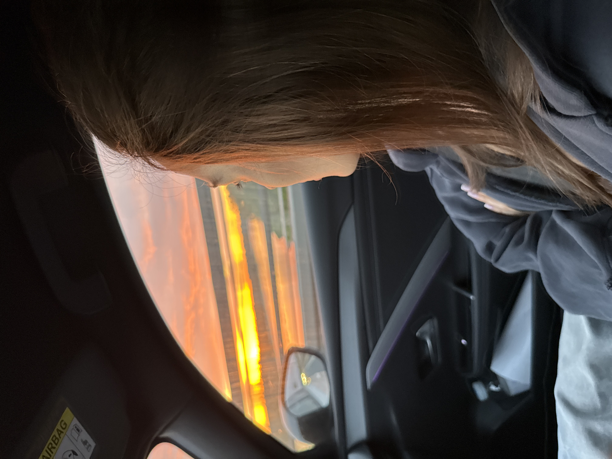 A side view shows a person with brown hair and a gray hooded sweatshirt. They are inside a car with a black interior. Outside the car window, there are vertical lines of orange and yellow light. A rearview mirror is visible. A yellow airbag warning label is visible.