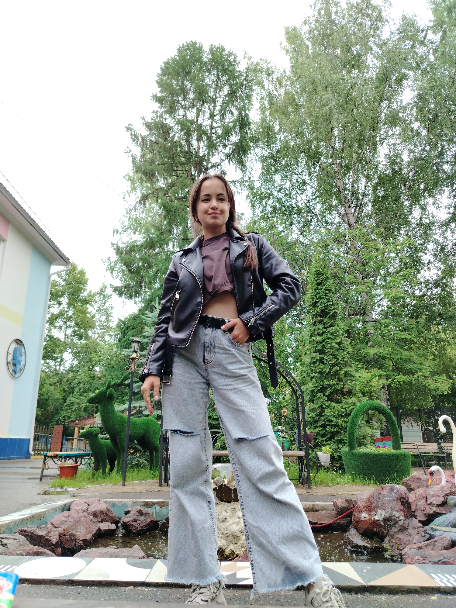 A young woman stands centrally, posing for a photo in an outdoor setting. She wears a black leather jacket over a dark, possibly brown or dark purple, cropped top, light-wash wide-leg ripped jeans, and light-colored sneakers. Her dark hair is styled with a braid visible on her left side, and she has a nose piercing. Her left hand is in her jeans pocket, and her right hand rests on her hip, with a ring visible on her right index finger. She looks slightly to her left with a gentle smile.
In the immediate foreground, there is a shallow decorative pool or fountain with reddish-brown rocks and some water, bordered by white, black, and grey geometric tiles. Behind the woman, on the left, are two green topiaries shaped like deer, one larger and one smaller. A black ornamental metal archway is visible to her right, along with a tall, slender green conifer. Further to the right, another green topiary in the shape of a basket with flowers and a white statue resembling a swan or goose are present. A black lamp post with an old-fashioned lantern is situated behind the left deer topiary. Wooden benches are partially visible in the midground behind the various garden features.
The background is filled with dense green foliage from numerous trees, including both deciduous and coniferous types. On the far left, a multi-colored building with light blue, light pink, and yellow stripes is visible, featuring a circular blue emblem on its wall. The sky is bright and overcast, indicating daytime. The setting appears to be a public park, garden, or recreational area, possibly associated with a community building in Chaikovsky, Russia. No visible text is present.
