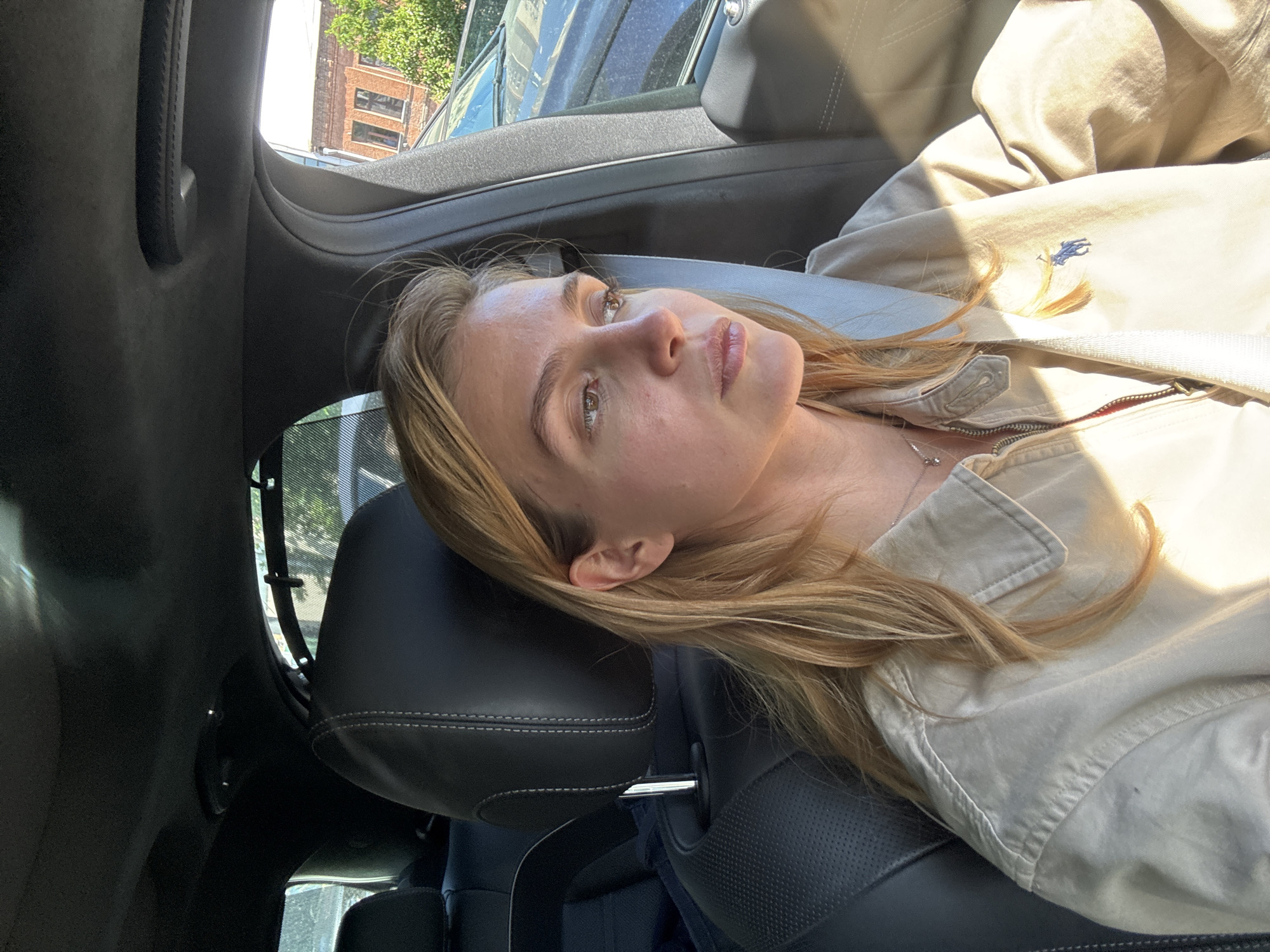 A young woman with long, light brown hair is depicted lying down, presumably in a car, looking upwards and slightly to her right. Her head rests on a dark, stitched leather car headrest. Strong sunlight illuminates her face, highlighting her forehead and nose, and casting bright patches on her hair and clothing.
She is wearing a light-colored, possibly beige or khaki, jacket or collared shirt. A small, embroidered blue logo, resembling the Ralph Lauren polo player, is visible on the upper right side of her garment. A thin silver necklace with a small charm hangs around her neck.
The setting appears to be the interior of a car, characterized by dark gray or black upholstery and trim. Through the car windows, elements of an outdoor urban or suburban environment are visible, including green trees, part of a brick building, and what looks like another vehicle. Daylight streams into the car, indicating it is daytime. Her expression appears calm and contemplative.