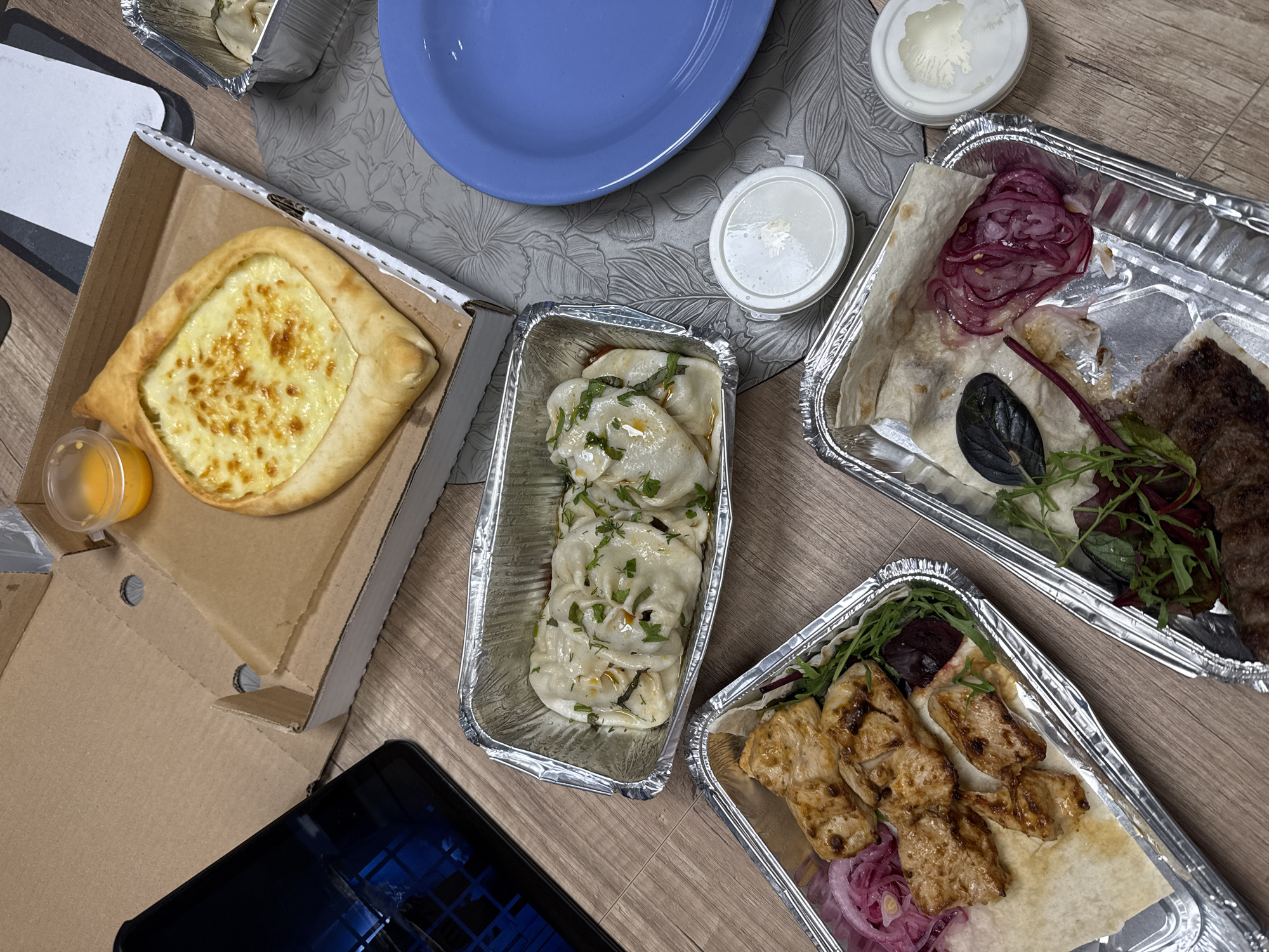 A top-down, medium shot captures a variety of takeout food containers arranged on a wooden surface. In the center, a boat-shaped pastry filled with melted cheese, likely Khachapuri, sits open in a cardboard pizza box, accompanied by a small plastic cup of orange sauce. To the left of the pastry, a smartphone with a reflective screen is partially visible. Below the pastry box, a rectangular aluminum foil container holds several portions of what appear to be dumplings or pierogi, garnished with chopped herbs. To the left of the dumplings, another aluminum container features grilled chicken skewers served with sliced red onion and a dark red vegetable slice. In the lower right quadrant, a third aluminum container contains pieces of flatbread with sliced red onion and a garnish of mixed greens. Two small white lidded containers, likely for dipping sauces, are also present. A blue plate is situated to the right of the food containers. The overall scene suggests a casual meal, possibly a takeout order, spread out on a table. The lighting is even and there are no visible people in the image.