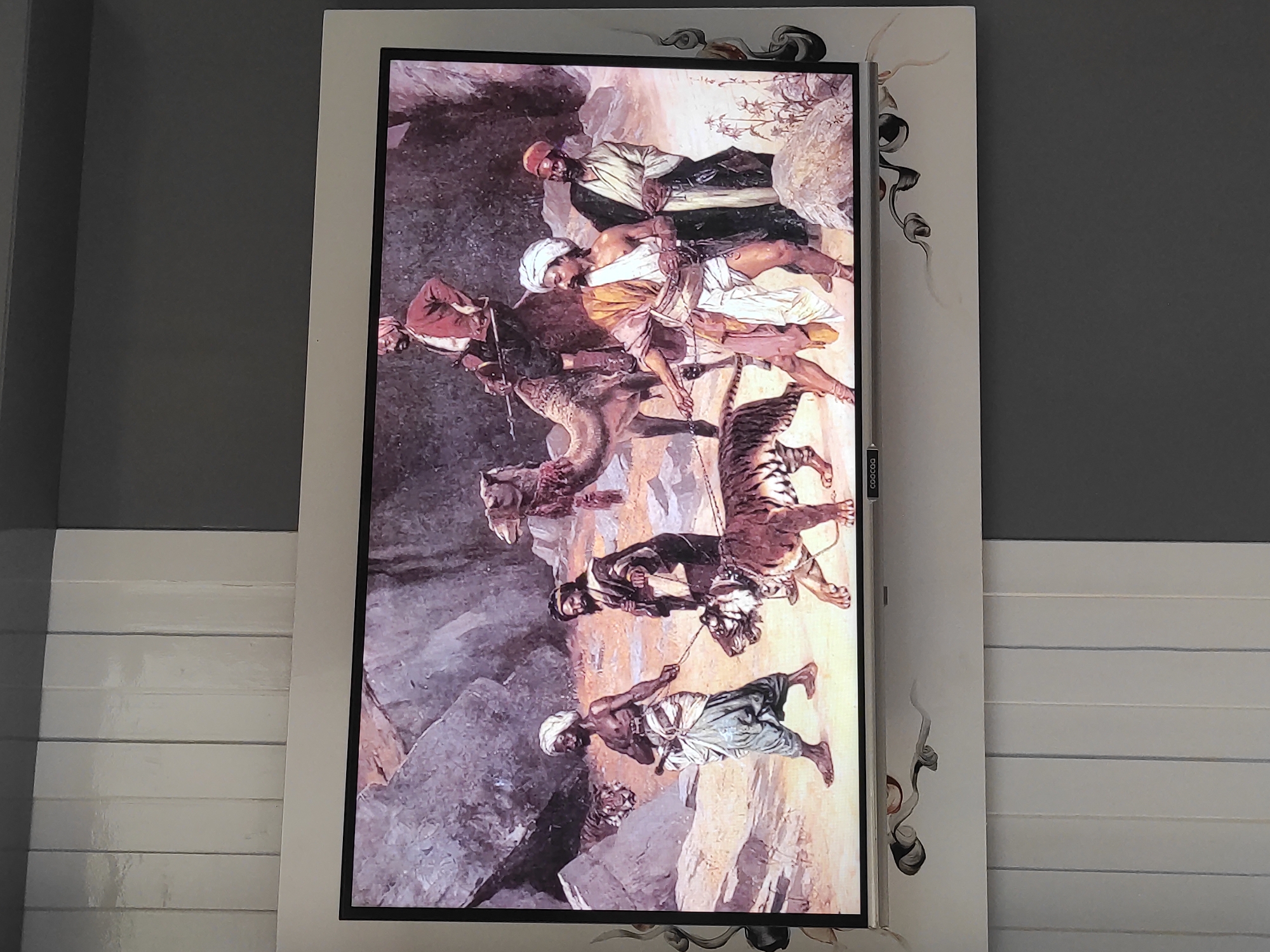 The image displays a painting on a television screen, depicting a scene with several individuals and animals in a rocky, arid environment. In the foreground, two men are walking, each leading a tiger on a leash. The man on the left is bare-chested, wearing a white turban and loose pants, and has a sword at his waist. The man on the right is wearing a turban and a robe. Behind them, a camel is visible with a rider on its back, who is holding a spear. Another tiger is partially visible in the background, near a rocky outcrop. The overall style of the painting suggests a historical or exotic setting. The television is mounted on a wall, with a decorative border around it, and the background of the room appears to be a wall with vertical white panels and a grey section above. The location in Sumedang, Indonesia is not evident from the image itself.
