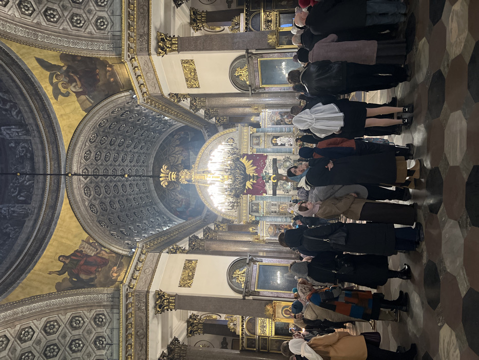 The image depicts the interior of the Kazan Cathedral in Saint Petersburg, Russia, viewed from the nave towards the iconostasis and main altar. The grand architectural space is characterized by massive, fluted columns with gilded Corinthian capitals, supporting high, vaulted ceilings adorned with intricate hexagonal patterns and gold accents. Large archways feature frescoes depicting religious figures, including Jesus with an outstretched arm on the left and angels on the right. A prominent, multi-tiered gilded chandelier hangs centrally, illuminated, before the elaborately decorated iconostasis. The iconostasis itself displays religious artwork, a large crucifix, and a purple banner with a golden sunburst emblem. The floor consists of hexagonal tiles arranged in a geometric pattern of light and dark brown shades. Approximately 30 to 40 individuals are visible, mostly adults, standing in groups and generally facing the iconostasis. Many women in the crowd wear head coverings, consistent with Orthodox Christian tradition. People are observed standing still, suggesting participation in or observation of a religious service. The scene is illuminated by the interior lighting and the chandelier, creating a solemn atmosphere.