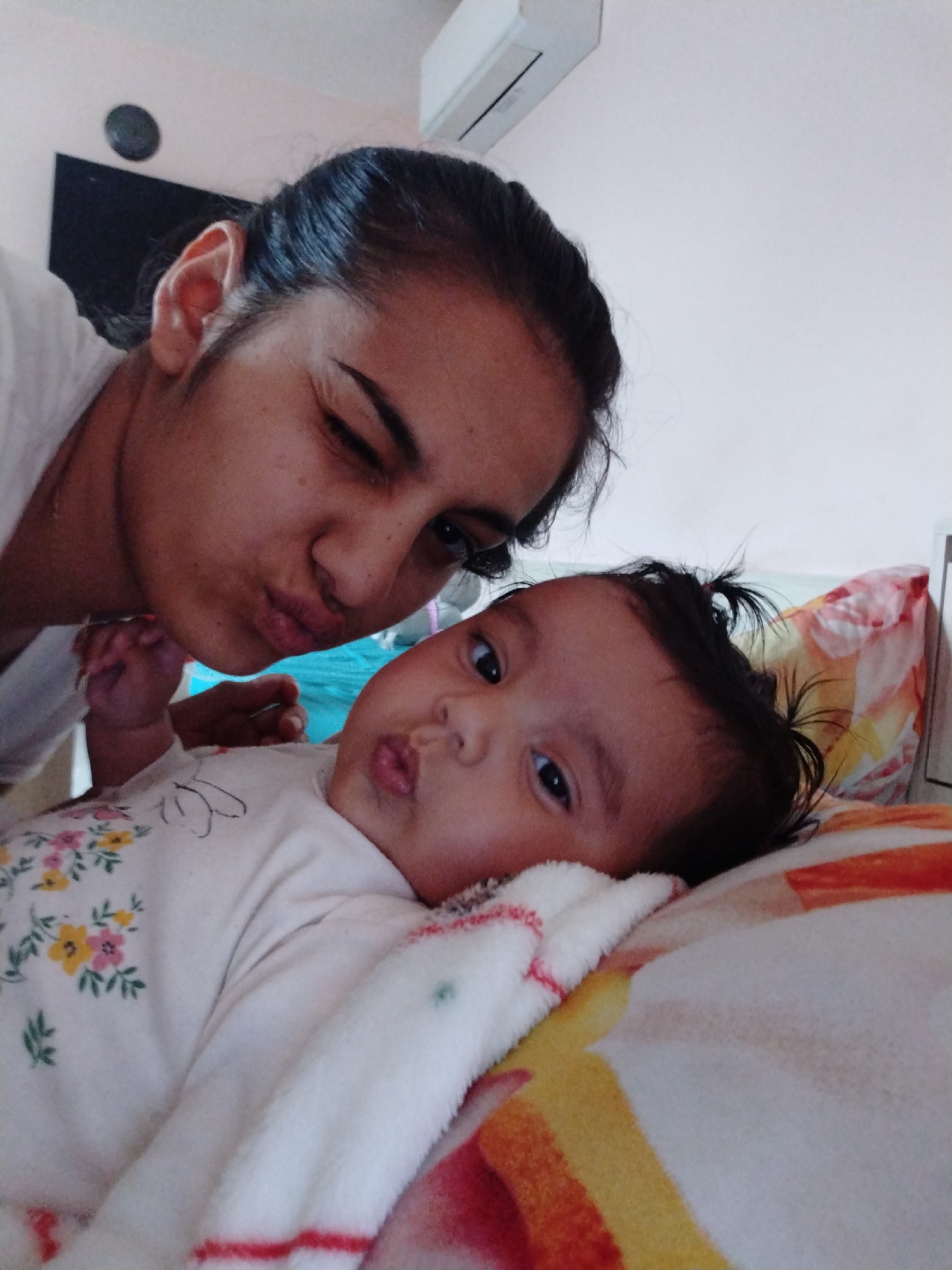 The image shows a woman and a baby in an indoor setting. The woman is positioned behind the baby, close to the camera. She is winking with one eye closed and her lips pursed as if giving a kiss. The baby is lying down on a patterned blanket or pillow, looking at the camera with a similar pursed lip expression. The baby is wearing a white outfit with a floral pattern on the upper part. In the background, there is a white wall and what appears to be an air conditioning unit. A dark-colored object, possibly a television, is visible in the upper left corner. Location is Unknown, Unknown.