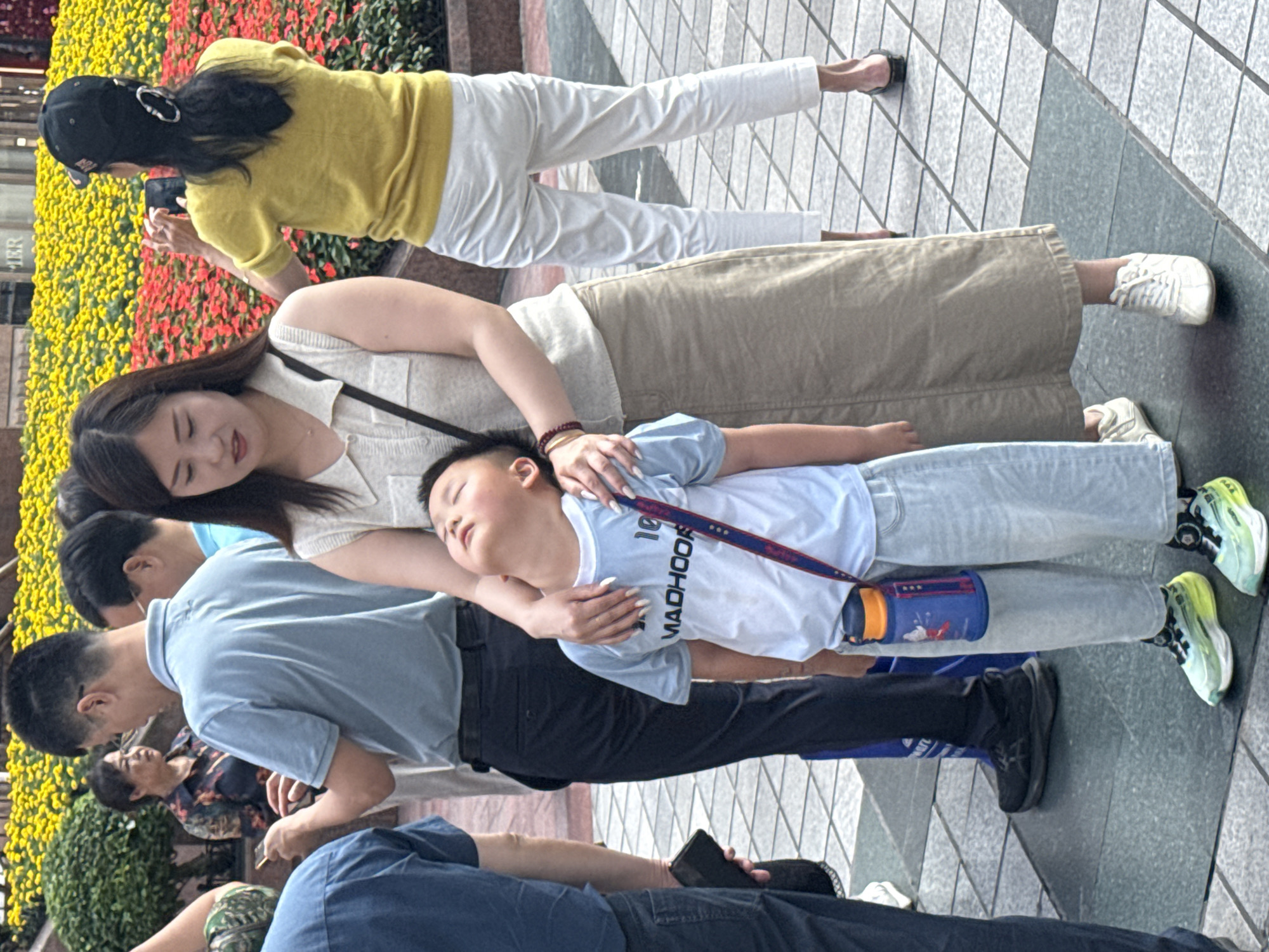 This image captures a vibrant daytime scene in what appears to be a public outdoor area, likely in Yuzhong District, China. The ground is paved with gray and black patterned tiles.
In the central foreground, a woman in a cream-colored top and a beige skirt gently supports a young boy, who appears to be sleeping or resting on her arm. The boy is dressed in a light blue t-shirt with the partial text "MA DHOO R" visible, light blue jeans, and colorful neon green and yellow sneakers. A blue and orange water bottle hangs from a strap around his neck. The woman's long, dark hair cascades down her back, and she wears a bracelet on her wrist. Her nails are long and light-colored.
Behind this pair, another woman, wearing a bright yellow long-sleeved top, white pants, and a black baseball cap, is actively engaged in taking a photograph with her mobile phone. She is facing a large display of lush flower beds filled with both yellow and red blossoms.
Several other individuals are present in the scene. A man in a light blue polo shirt and dark pants stands near the woman and boy. Another man, wearing a dark blue shirt and pants, is partially visible in the lower right, holding a mobile phone. Various types of footwear are visible on different subjects, including white sneakers and black sandals.
In the background, beyond the flower beds, a building with indistinct features shows partial text that appears to read "TER." The overall atmosphere suggests a casual public outing or leisure activity.