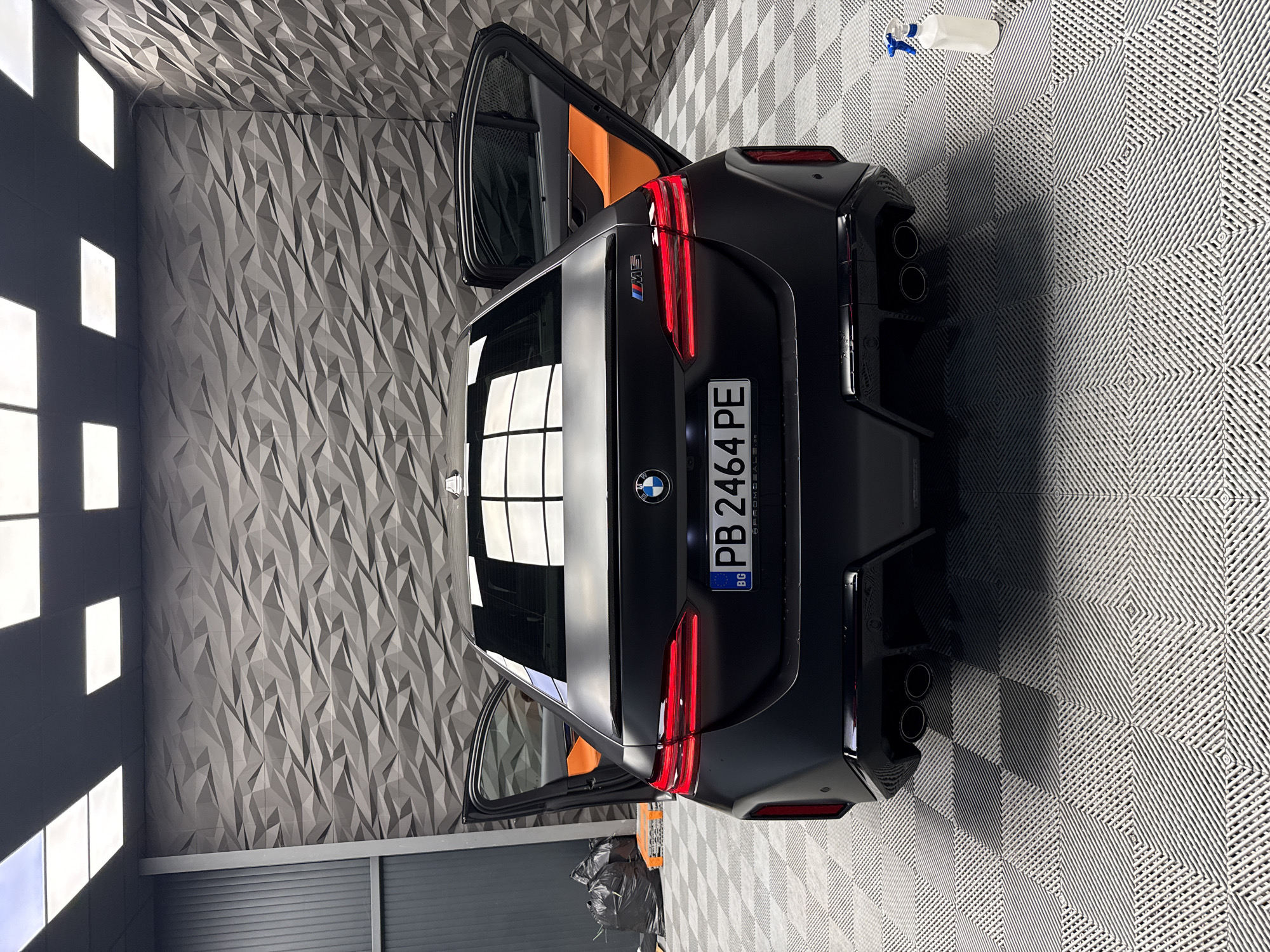 A vertically-oriented image shows the rear of a matte black BMW M5 CS car inside what appears to be an auto detailing or showroom space. The car is parked on a grey and white checkered floor. The rear doors of the car are open, revealing orange interior panels. The car's license plate reads "PB 2464 PE" with a "BG" abbreviation in the lower left, indicating Bulgaria.
The room features a wall with a geometric triangular texture. To the left, a series of square light fixtures are visible on the wall. In the top right corner, a white spray bottle sits on the floor. A trash bag is visible beneath one of the open doors.
The license plate indicates that the car is registered in Bulgaria, but there is no way to confirm it is in Plovdiv.