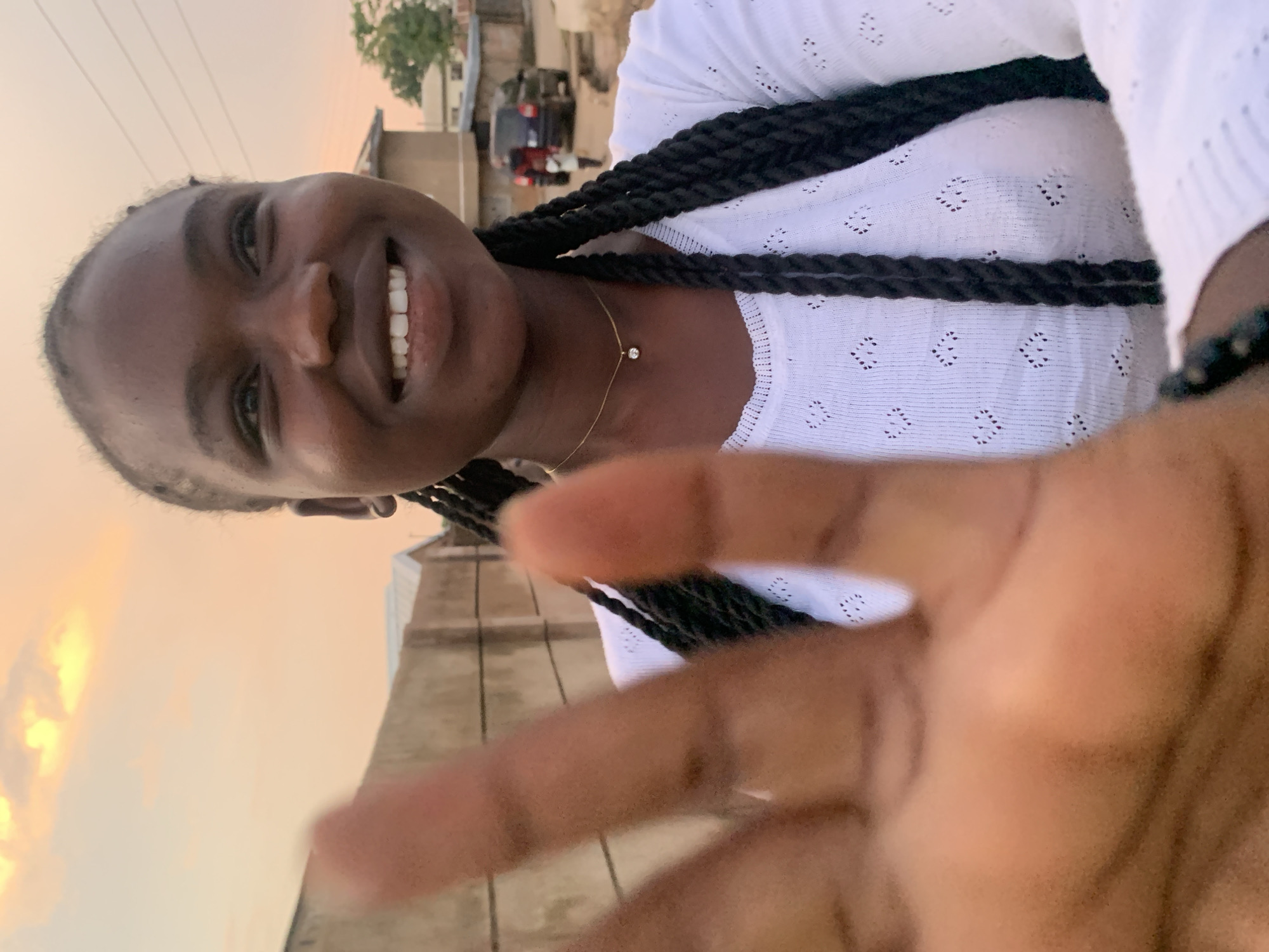In a close-up, outdoor selfie taken during golden hour in Jos, Nigeria, a young Black woman with braided hair smiles warmly. She wears a white, textured long-sleeved top and a delicate gold necklace with a single pendant. Her right hand is blurred in the foreground, obscuring part of the frame. The background features a concrete wall, a building, a car, and a person walking in the distance. The sky is illuminated by the warm hues of the setting sun.
