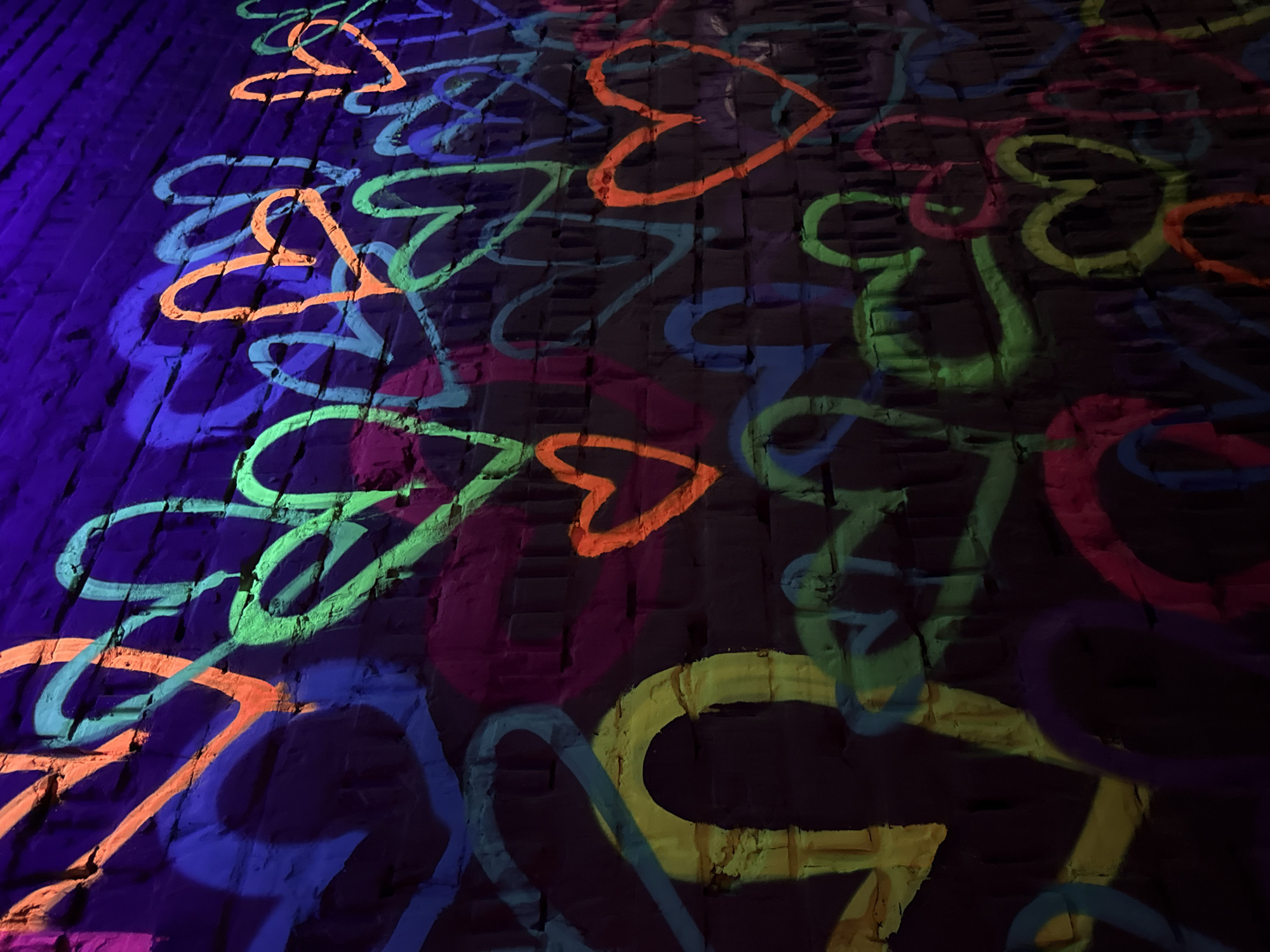 This close-up image showcases a vibrant, glowing artwork displayed on a dark, textured surface, likely a brick wall or pavement. The scene is illuminated by ultraviolet (UV) light, causing various heart shapes and abstract, swirling lines to fluoresce brightly against the deep, dark blue and purple background.
The artwork features numerous hearts of different sizes and orientations, along with graffiti-style wavy lines. The colors of the glowing elements include neon green, bright orange, light blue, yellow, pink, and purple, creating a dynamic and energetic visual. The shapes are scattered across the surface, occasionally overlapping, adding to the layered effect.
The texture of the underlying surface is visible beneath the glowing paint, indicating an uneven, brick-like material. The dark ambient lighting suggests a nighttime setting or a dimly lit interior space. There are no people or specific activities depicted, focusing solely on the artistic display. No visible text is present in the image. The location is Moscow, Russia.