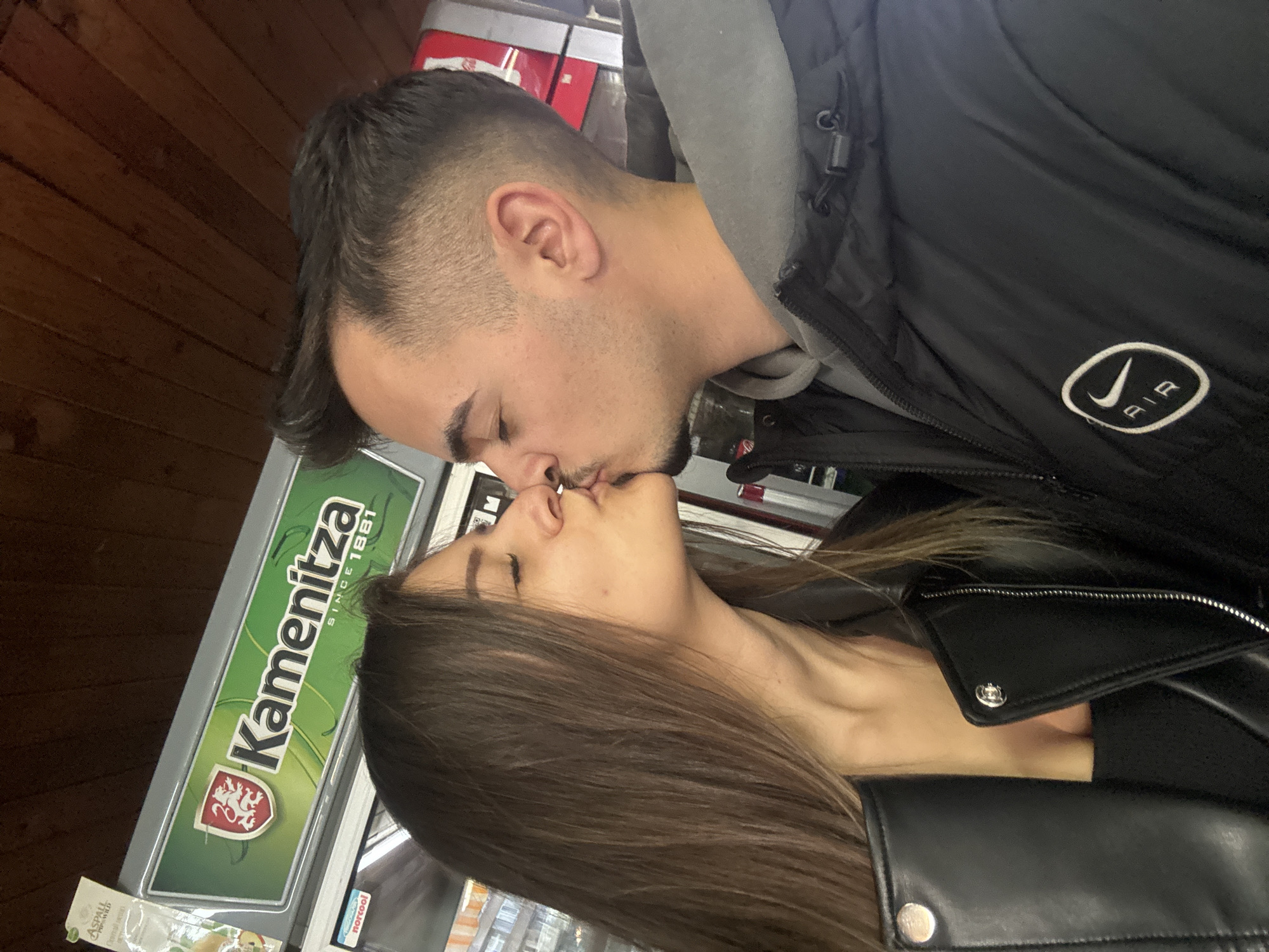A man and woman are kissing. The man is wearing a black Nike Air jacket and a gray hooded sweatshirt. The woman is wearing a black leather jacket. In the background is an advertisement for Kamenitza beer. The location is Sofia, Bulgaria.