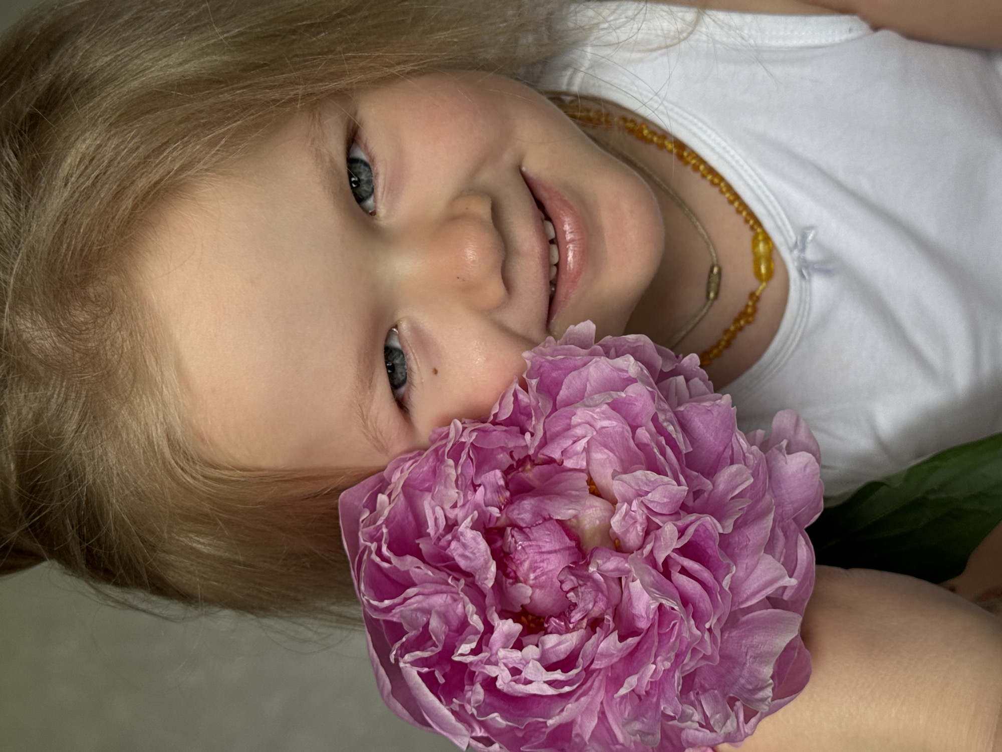 In Ферма, Russia, a fair-skinned young child with blonde hair and blue eyes is positioned in a close-up, chest-up shot against an out-of-focus, light gray textured background. The child is wearing a white sleeveless top with a small white bow detail near the neckline. Two necklaces are visible: a shorter, thin gold-colored chain with an oval bead, and a longer strand of amber-colored beads. A large, fully bloomed, multi-petaled peony flower, predominantly fuchsia/magenta in color, is held by the child's left hand (viewer's left) close to the left side of her face, partially obscuring it. A green leaf is visible at the base of the flower. The child is looking directly forward, exhibiting a slight smile that reveals her upper front teeth. A small, dark mole is present on the skin below her left eye (viewer's left).