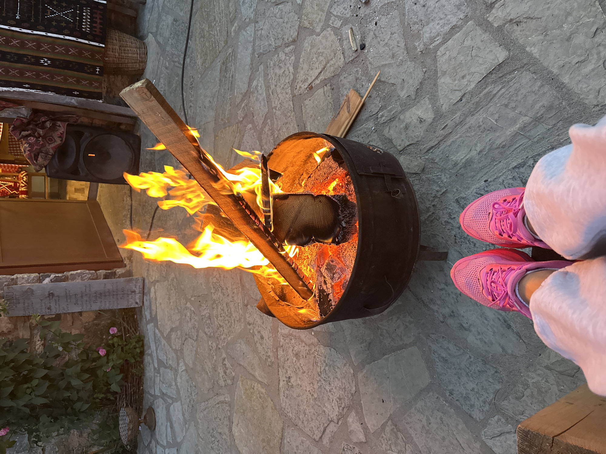 The image captures a warm outdoor scene focused on a crackling bonfire in a metal fire pit. The ground is paved with large, irregular flagstones. In the foreground, a person's legs are visible, wearing bright pink sneakers and light-colored pants, suggesting they are seated and enjoying the fire.
The fire pit is filled with burning logs and embers, with flames leaping upwards. A piece of wood extends out of the fire pit, angled towards the viewer. In the background, to the left, is a stone wall covered in greenery and some pink roses, with a rustic wooden sign that appears to be a menu. Further back, to the right, is a dark-colored speaker and some decorative woven textiles hanging on a wall, with a wicker basket nearby. The overall ambiance is cozy and inviting.
The image was taken in Gusinje, Montenegro, a town known for its natural beauty and traditional architecture. The setting appears to be an outdoor area, possibly a courtyard or patio of a restaurant or guesthouse, given the presence of a menu and the rustic decor. The lighting suggests it is either dusk or early evening.