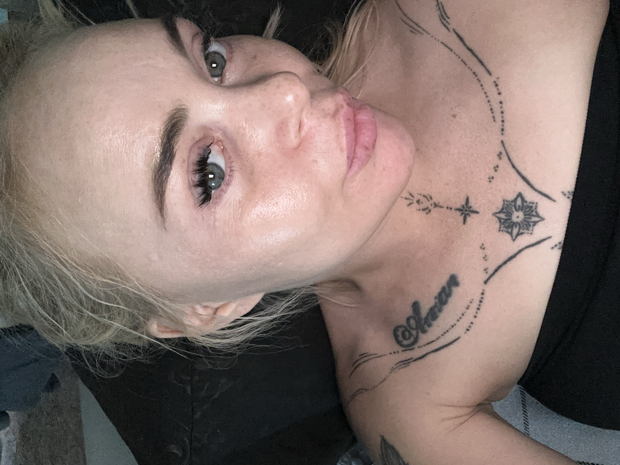 The image is a close-up portrait of a fair-skinned female with blonde hair, seen from the chest up. Her hair is pulled back, with several wispy strands framing her face. She has blue-grey eyes with prominent dark eyelashes and dark, well-defined eyebrows. Her nose is straight, and her light pink lips are slightly pursed. A visible sheen is present on her forehead and cheeks. She is wearing a black strapless top or bandeau. Multiple black ink tattoos are visible on her upper chest and collarbone area: on her left side, a script tattoo reads "@Aerian," and across her décolletage, there are various geometric and dotted line patterns, including a four-pointed star, a seven-pointed starburst, and a floral mandala design. A small, stylized leaf or flower symbol is also visible near her throat, connected by a dotted line to a smaller star. The subject is looking directly into the camera. The background is dark and indistinct, suggesting an indoor setting, with a portion of light blue patterned fabric visible on the lower left. The location is unidentifiable.