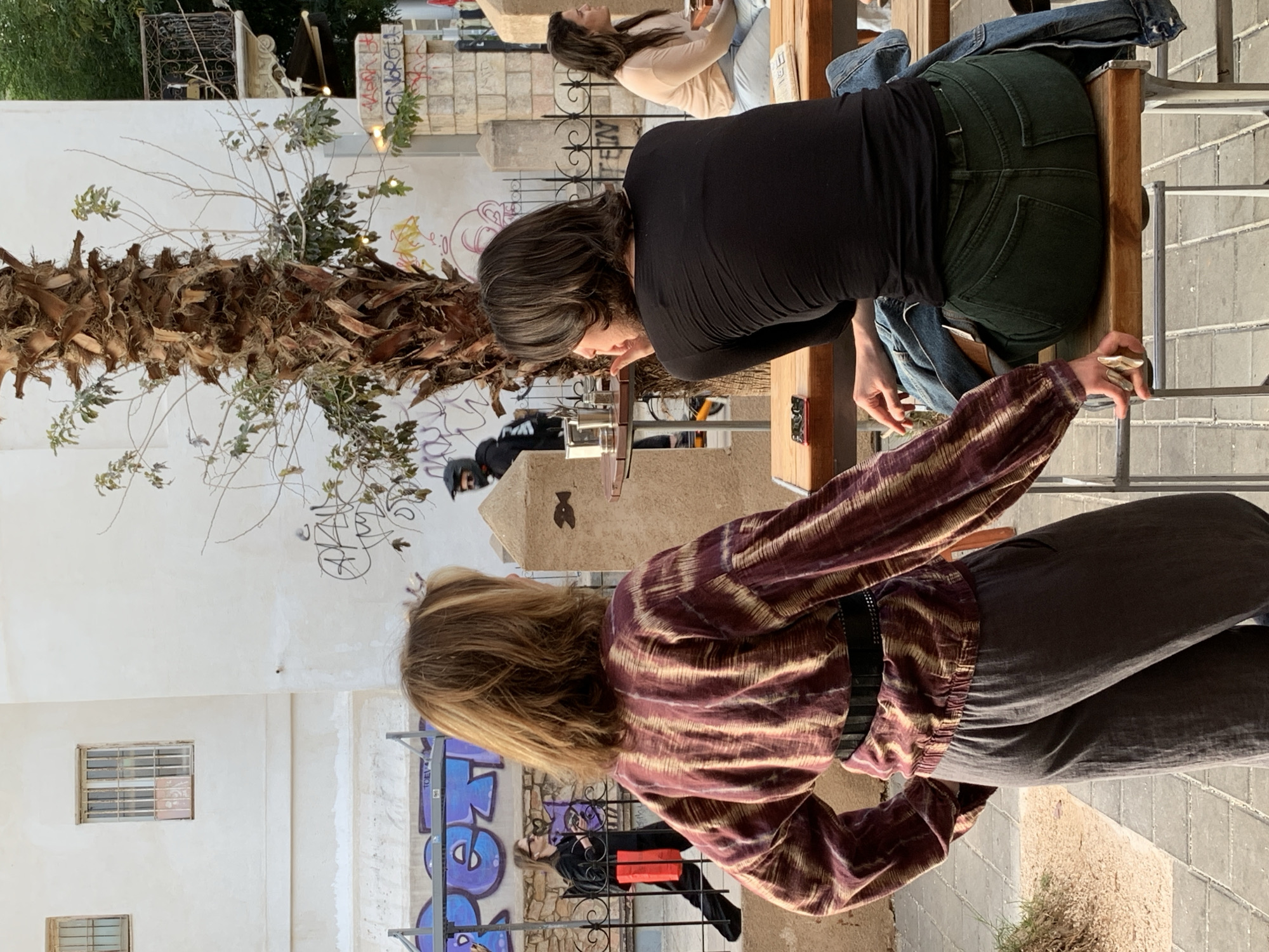 This image captures a casual outdoor scene at a cafe or restaurant, likely in Tel-Aviv, Israel, on a bright, sunny day. The perspective is slightly elevated, looking down into the bustling area.
In the foreground, a woman with shoulder-length blonde hair is seen from behind and slightly to the side. She wears a striking patterned long-sleeved shirt in shades of reddish-brown and beige, paired with dark grey trousers and a black belt. Her right hand is extended forward, holding a small, light-colored object.
Further into the scene, a second woman with dark, shoulder-length hair is seated at a light wooden table, her back mostly to the viewer. She is dressed in a black long-sleeved top and dark green jeans, with a denim jacket draped over her chair. She appears to be reaching for something on the table, where a small red-backed phone is visible. A third woman with lighter hair is seated at another table in the background, seen in profile.
The setting features white buildings with small, barred windows. A prominent palm tree with a rough, textured trunk and sparse branches dominates the left side of the frame. The ground is paved with light-colored tiles. Graffiti, including purple and black letters and designs, is visible on the walls in the background. One piece of graffiti appears to spell "GNORGU" in white letters on a dark surface. In the distance, a person wearing a dark jacket and helmet is partially visible behind the palm tree. The overall ambiance is relaxed and casual, suggesting people enjoying refreshments outdoors.