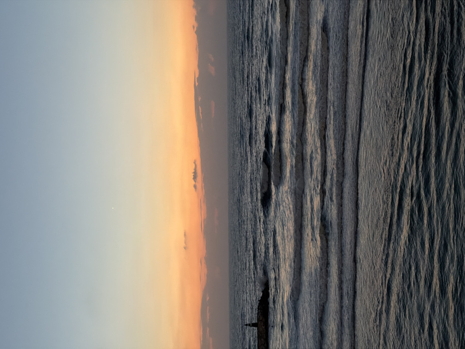 The image depicts a sunset over the Mediterranean Sea along the coastline of Tel-Aviv, Israel. The sky transitions from light blue at the top to a vibrant orange and yellow hue near the horizon, where scattered dark clouds are visible. A single, bright celestial object, possibly a star or planet, is present in the upper-left quadrant of the sky. The sea occupies the lower two-thirds of the frame, exhibiting a dark blue-grey color with numerous parallel waves rolling towards the foreground, displaying white crests where they break. On the far left, a dark, conical lighthouse stands atop a rocky breakwater that extends into the sea from the left edge of the frame. Waves are observed impacting against this breakwater. No individuals or vessels are visible. The scene is dominated by natural elements, indicating dusk or late evening conditions.
