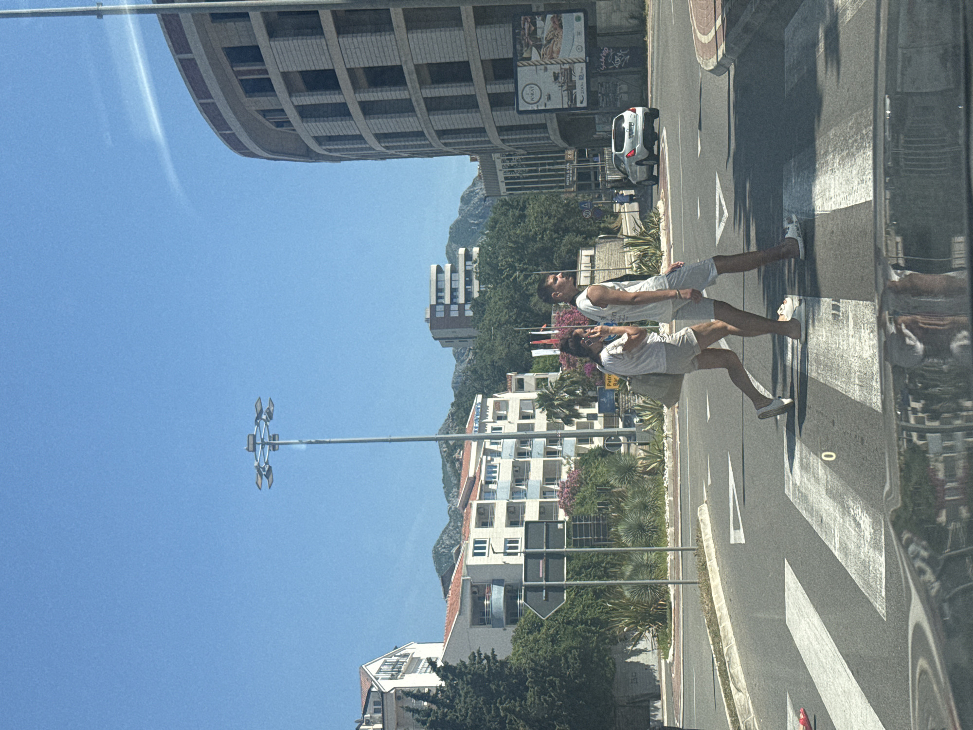 The image depicts a sunlit street scene in Rafailovići, Montenegro, captured from the interior of a vehicle, evidenced by a reflection on the lower edge. The sky is clear and blue.
In the foreground, a marked pedestrian crossing spans an asphalt road. Two individuals are traversing it from left to right. The person on the left, a woman, wears white shorts, a white top, and white slip-on sandals, and carries a light-colored shoulder bag. She is holding a phone to her right ear. The person on the right, a man, wears light-colored shorts, a white tank top, and white slip-on sandals. He looks towards the woman.
Immediately beyond the pedestrians, a grey directional arrow sign on a pole is visible, pointing left. A central median strip with green vegetation, including succulents and small trees, separates the road. Further down the road on the right, a white SUV is stopped or parked. A circular speed limit sign displaying "50" is partially visible near the SUV.
On the left side of the road, multi-story white buildings with red tiled roofs and blue balconies are partially obscured by dense green foliage, including cypress and palm trees. Mountains are visible in the distant background beyond these structures.
On the right side of the road, a prominent, curved, multi-story building with a textured facade of rectangular panels dominates the midground. Dark windows are interspersed across its surface, and the upper section features a reddish-brown band. A large billboard displaying "POBJAT" and food imagery is affixed to this building. Below the billboard, graffiti is present on a wall. Another building with multiple balconies is visible behind the curved structure. A sign partially reading "HOTEL & SPA" is also visible. A tall, multi-headed street light pole stands centrally in the upper part of the image. A yellow sign reading "Petrovac Bar" is visible on the far right. Distant mountains rise behind the buildings on the right.