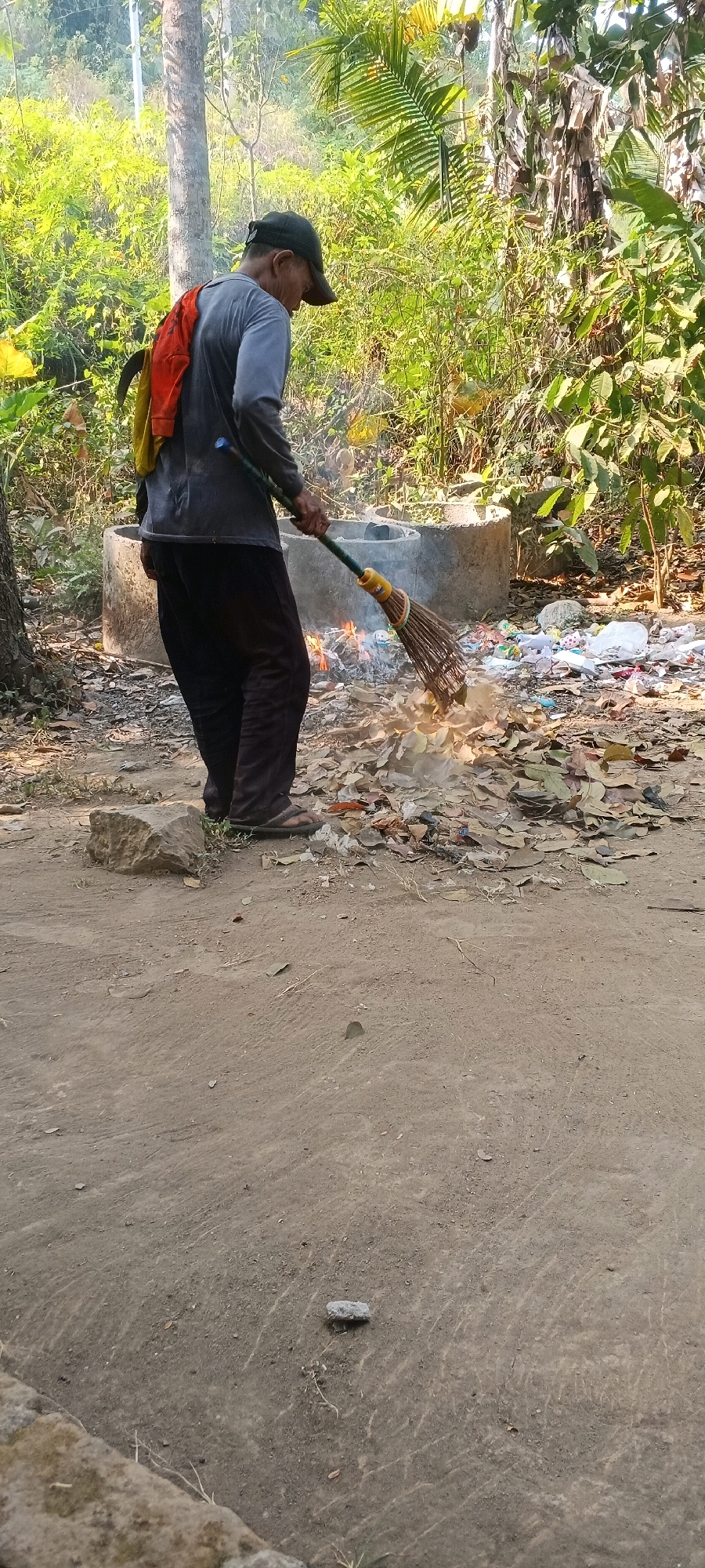 An elderly man, wearing a grey long-sleeved shirt, dark pants, sandals, and a dark baseball cap, is burning a pile of dry leaves and debris. He is in the process of sweeping more leaves into the fire with a broom that has a green handle and a straw brush. The fire is in a concrete ring, and behind it, another identical concrete ring is visible. Scattered around the burning area are various pieces of trash, including white and colored plastic bags and wrappers. The ground is dirt, with scattered rocks and dry vegetation. Lush green foliage and trees form the background, suggesting an outdoor, possibly semi-rural, location. The lighting indicates it is daytime.