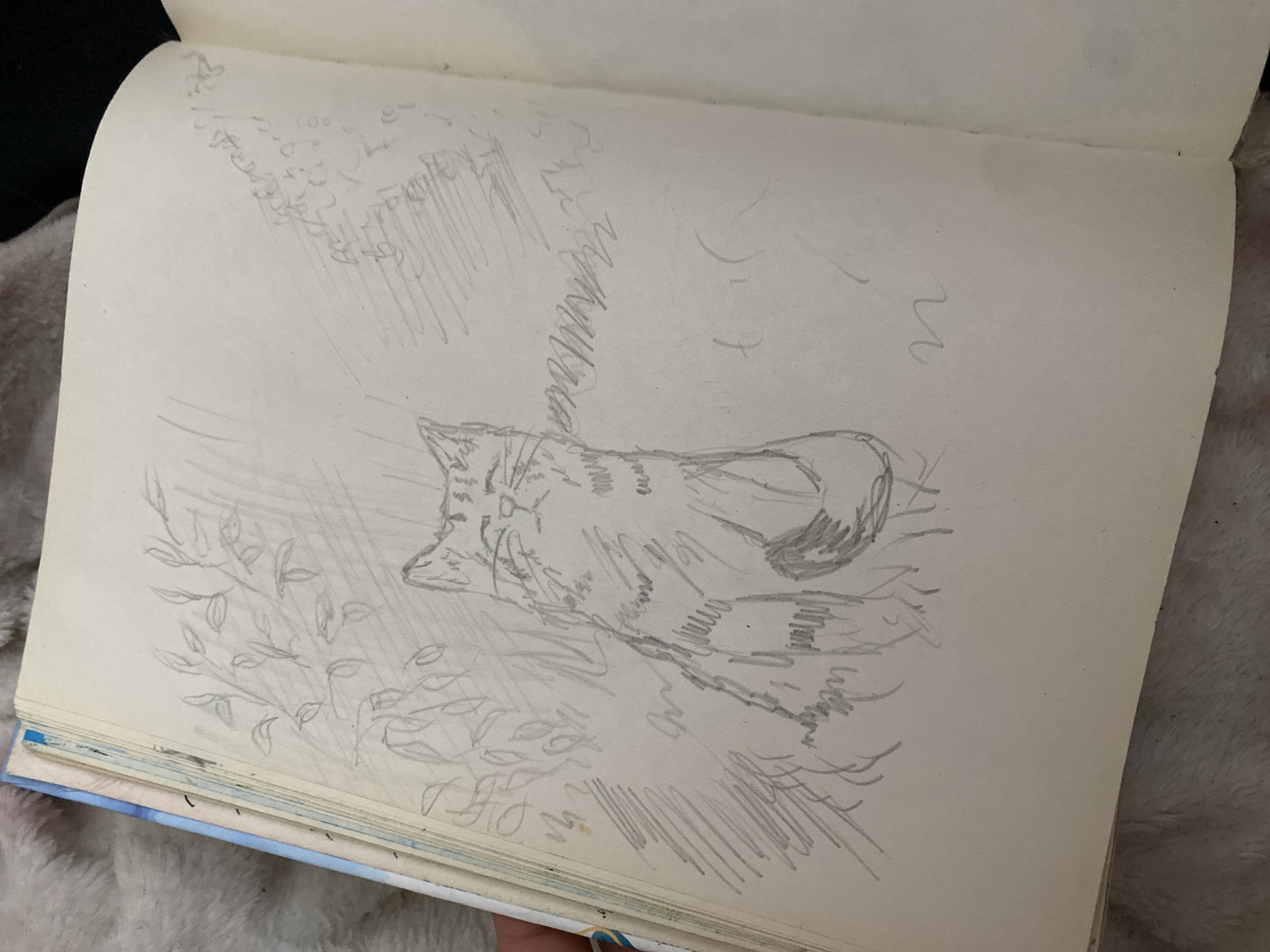 A pencil sketch on a notebook page depicts a cat lying down in a natural setting, possibly outdoors among foliage. The cat is the central subject, shown in profile with its head turned slightly towards the viewer. Its body is curled, and its tail is visible. The sketching style is loose and gestural, with quick lines suggesting fur texture and the form of the cat. Around the cat, there are faint sketches of leaves and branches, indicating it is resting amidst plants. There are also abstract lines and shading in the background, possibly representing grass, wind, or other environmental elements. The overall impression is of a relaxed moment captured in a quick drawing. The notebook is open, resting on a textured surface, and a hand is holding the bottom edge. There is no visible text in the image. The scene is undated and offers no specific time of day or weather cues beyond what might be inferred from an outdoor setting.