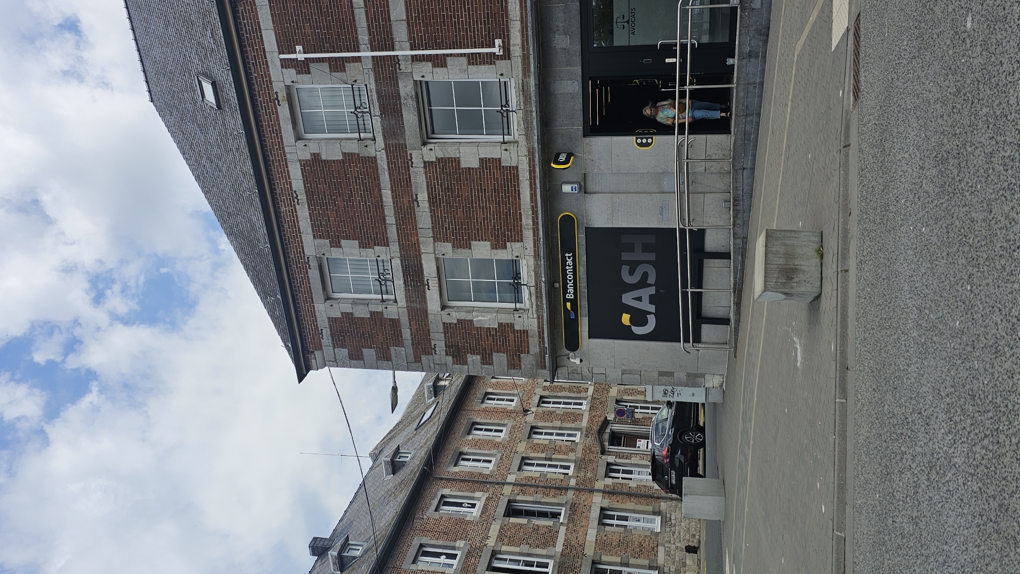 The image depicts a street view in Philippeville, Belgium. On the right side of the frame is a brick building with stone accents, housing a "CASH" establishment. A yellow sign with "Bancontact" is visible above the entrance. A woman is seen exiting the "CASH" establishment. To the left of this building, another brick building with multiple windows is partially visible, suggesting an urban or semi-urban environment. A dark-colored car is parked on the street in front of the buildings. The ground is paved with interlocking stones and asphalt. The sky is partly cloudy.