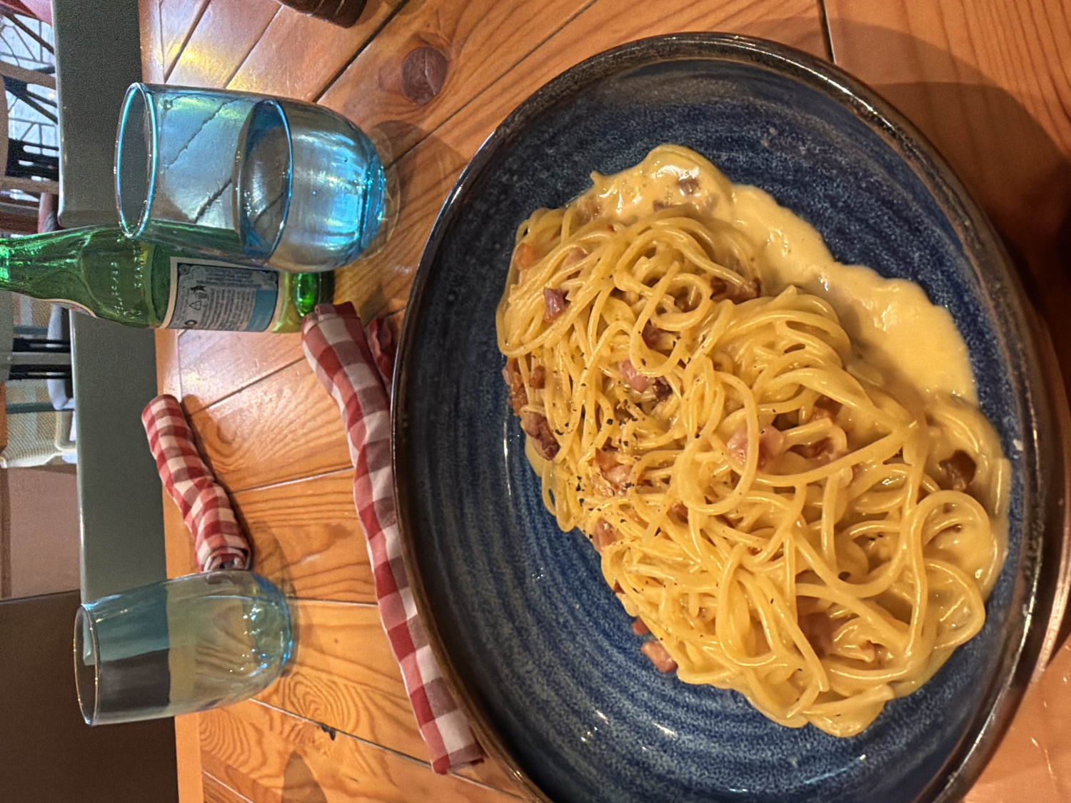 The image displays an overhead view of a wooden table set for a meal in an indoor dining establishment, likely a restaurant or cafe in Sofia, Bulgaria.
The central focus is a large, generous serving of spaghetti, identifiable as Carbonara, presented on a dark blue, textured ceramic plate. The pasta is coated in a creamy, rich yellow sauce, featuring small pieces of browned meat (such as guanciale or pancetta) and sprinkled with black pepper. The sauce appears thick and well-emulsified, pooling slightly at the bottom of the plate.
To the left of the plate, a green glass bottle with a white and blue label stands upright. The label clearly shows the words "ACQUA MINERALE," indicating it contains mineral water. Adjacent to the bottle, a translucent blue glass filled with water is placed, while a second, empty blue glass rests further down on the wooden table. Two red and white checkered cloth napkins, neatly rolled, are positioned on the wooden surface, one near each glass. The table itself is made of light-colored wood with a prominent grain.
The scene conveys a casual and welcoming atmosphere, suggesting a relaxed mealtime. No individuals are visible in the frame, and the time of day is not discernible from the indoor lighting. The image captures the setup for a meal, emphasizing the prepared dish and accompanying drinks.