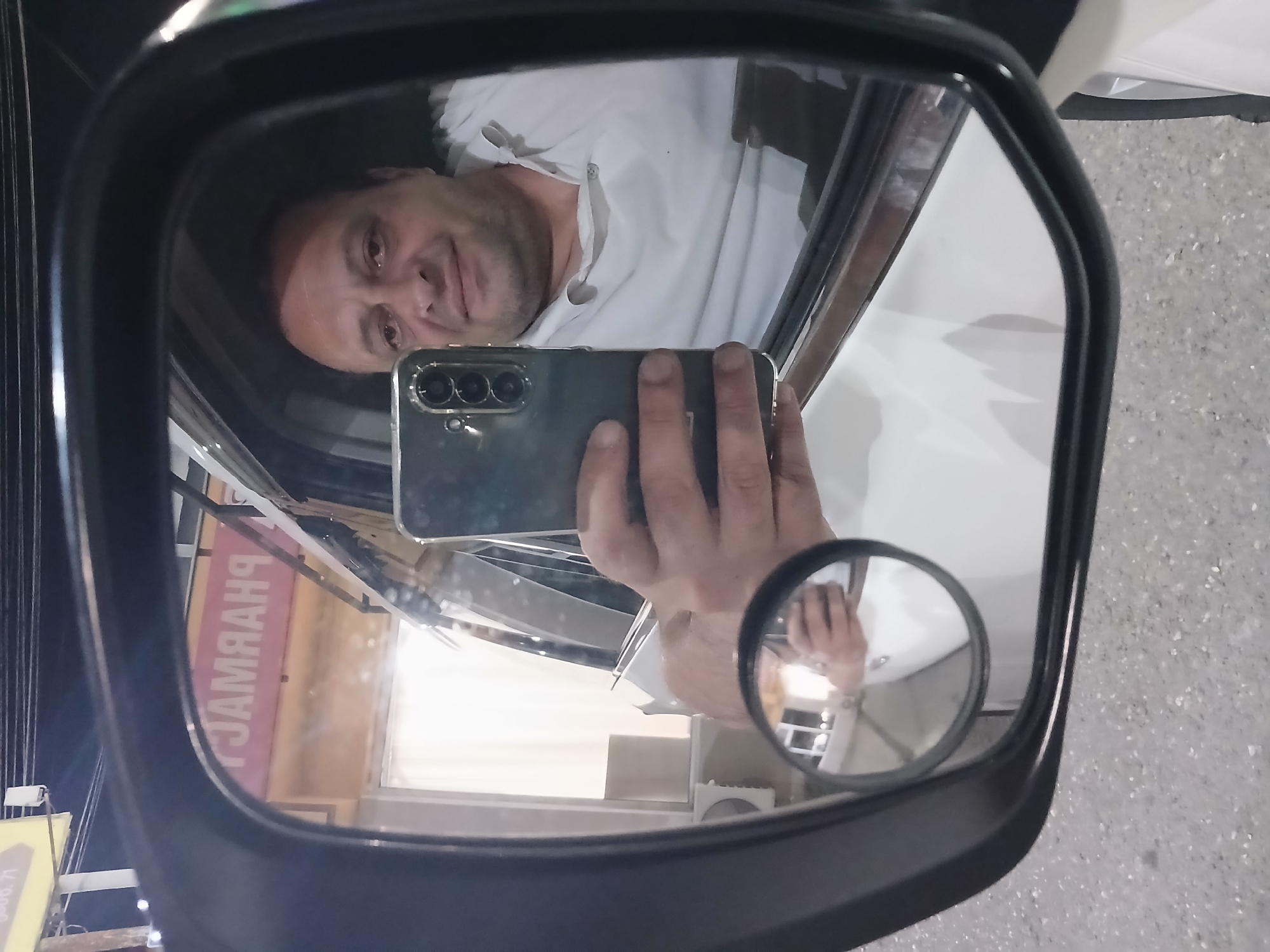In Telavi, Georgia, a man takes a selfie in the reflection of a car's side-view mirror. He is seated inside the vehicle, holding a smartphone with a triple-lens camera. The mirror also reflects a sign in the background that reads "PHARMACY" in pink lettering. The overall scene is captured at night, with artificial lighting illuminating the surroundings. The car appears to be parked on a paved surface.