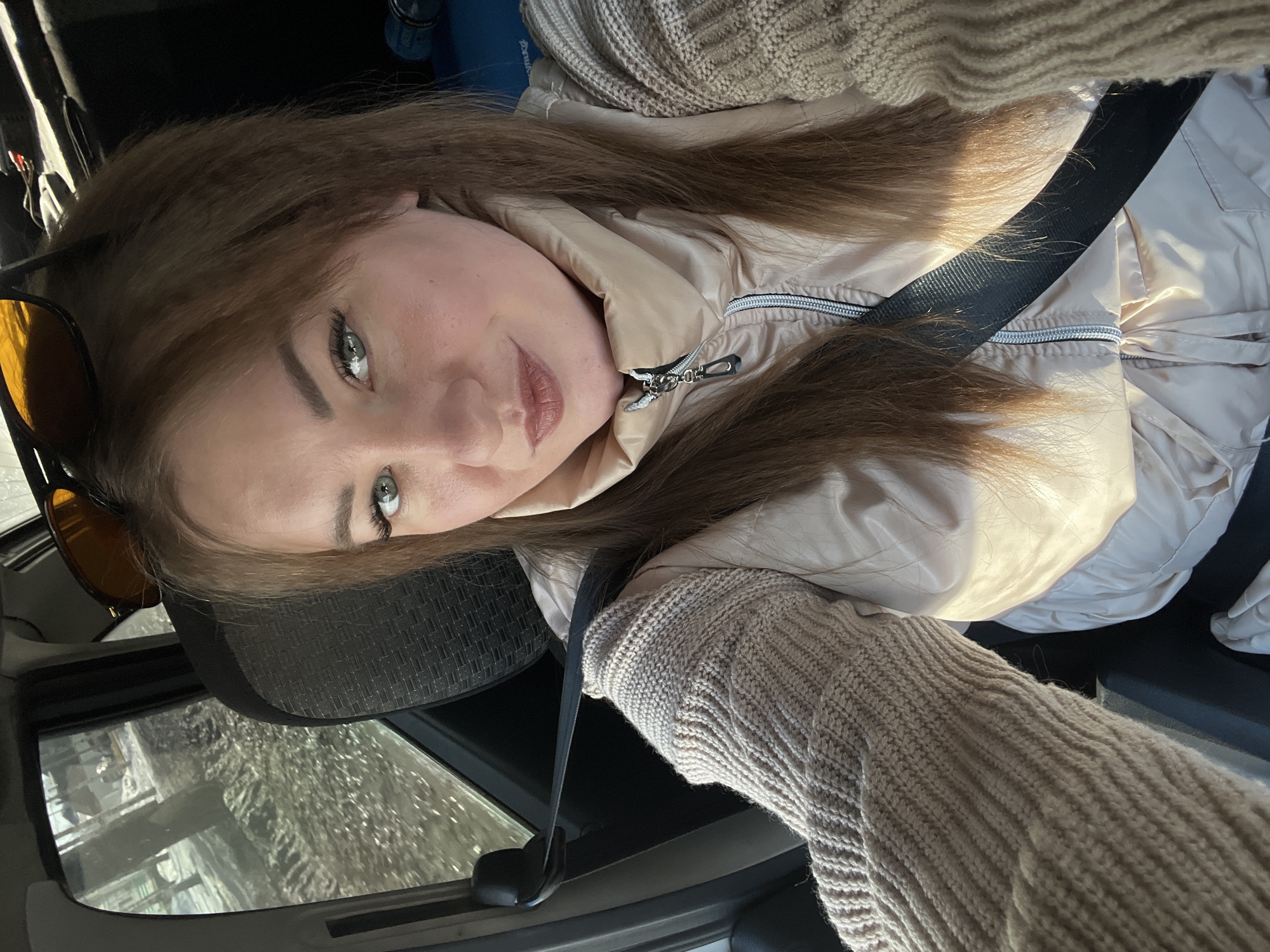 A selfie depicts a young woman with long, auburn hair and green eyes, wearing a tan-colored knitted sweater and a light beige jacket. She is wearing a seatbelt and sitting in a car. The car is driving through Chaikovsky, Russia. The passenger-side window is visible, and the background outside appears to be a winter scene with trees. Sunglasses are placed on her head. The time of day appears to be afternoon or early morning, judging by the light.