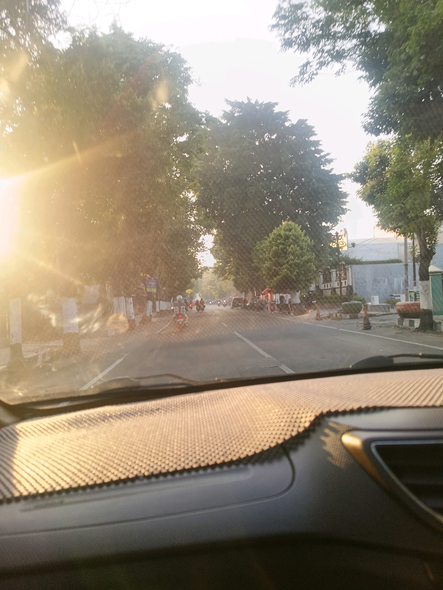 The image depicts a view from inside a vehicle, looking through the windshield at a street scene in what is identified as Jombok, Indonesia. The sun is low in the sky, casting a strong glare and lens flare across the left side of the frame, illuminating trees lining the road. The road appears to be a two-lane street with light traffic, primarily consisting of motorcycles. Several individuals are visible riding motorcycles, some with passengers. On the right side of the road, there are buildings and what appear to be some small vendor stalls or shops. The foreground shows the dashboard of the vehicle, with a textured surface in front of the windshield. The overall atmosphere is that of a sunny, possibly early morning or late afternoon street view.