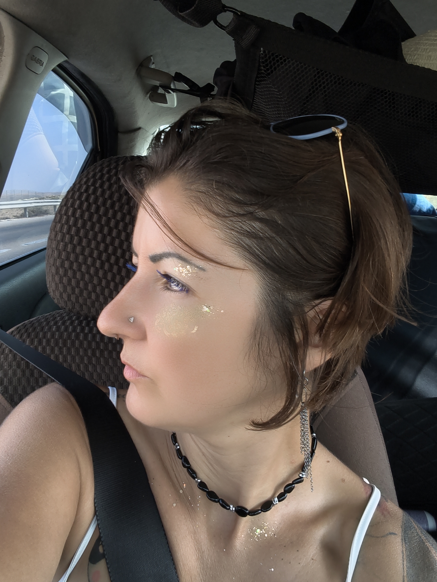 A profile of a woman's face and upper body is shown from the chest up, taken inside a vehicle. She is looking to her left, away from the camera, with her mouth slightly open. She has brown hair pulled back, and sunglasses resting on top of her head. She is wearing blue eyeliner and has glitter on her eyelids and cheeks. A silver nose stud is visible. She is wearing a black beaded necklace and a silver chain earring. Her left shoulder is visible, and she is wearing a white strap top. A black seatbelt is visible across her chest. The background shows the interior of a car, with a patterned seat and a window with a view of a road and landscape, suggesting the vehicle is in motion. The location is Ramat Negev Regional Council, Israel.