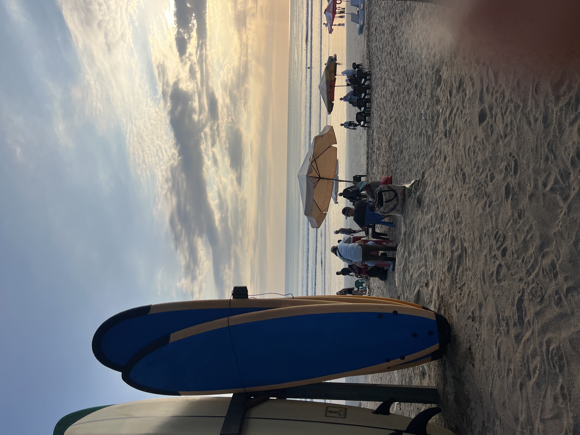 This image depicts a beach scene in Kuta, Indonesia, at dusk. In the foreground, two surfboards are propped upright on the sand, one with a white and green exterior, and the other a prominent blue with yellow accents. Beyond the surfboards, the beach is populated with several individuals and groups. Some people are seated under umbrellas or on lounge chairs near the water's edge, while others appear to be standing or walking. The ocean stretches to the horizon, with small waves rolling in. The sky is filled with a mix of clouds, illuminated by the setting sun, casting a warm, golden light over the scene. The overall atmosphere is one of relaxation and leisure, typical of a popular tourist destination like Kuta.