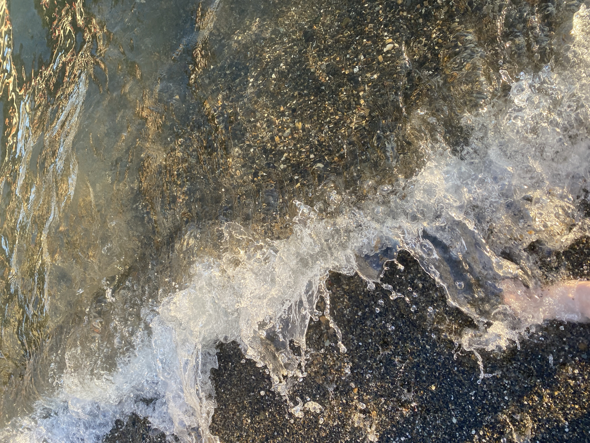 The image shows a close-up, overhead view of a wave breaking on a pebble beach. The water is clear and reflects sunlight, creating shimmering patterns on the wet pebbles. Small pebbles of various colors, including black, gray, and white, are visible beneath the shallow water. The wave is frothy and dynamic, with droplets of water suspended in the air, capturing a moment of movement. In the lower right portion of the image, a person's foot is partially submerged in the water and sand, suggesting a leisurely activity like wading or enjoying the sea. The scene evokes a sense of warmth and relaxation, likely during a sunny day. The lighting indicates it could be either morning or late afternoon due to the warm, golden hues reflecting on the water and pebbles. The location is identified as Sochi, Russia. There is no visible text in the image.