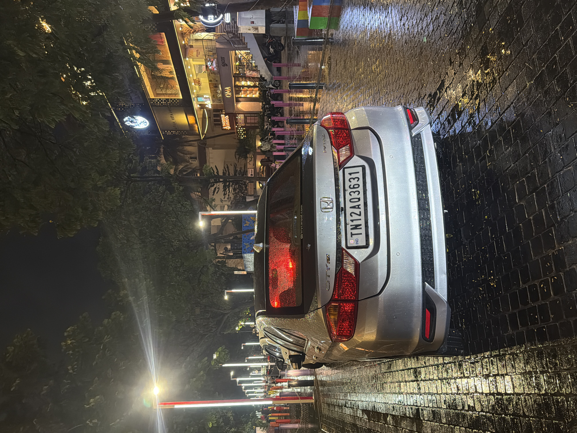 The image depicts a rear view of a silver Honda City sedan parked on a wet, cobblestone street at night. The car's brake lights are illuminated red, reflecting on the wet surface. A license plate with "TN12 A03631" is visible. To the left, a line of illuminated poles with red and white markings extends into the distance, indicating a pedestrian walkway or street demarcation. Trees line the street, and beyond them, a building with multiple illuminated floors is visible. The building's signage includes the words "OMA" and "Filli cafe". The scene suggests a commercial area in Chennai, India, possibly in the evening after rain. The lighting is atmospheric, with streetlights and building lights creating reflections on the wet ground. No people are clearly visible in the immediate vicinity of the car, but some figures can be faintly seen in the background near the building.