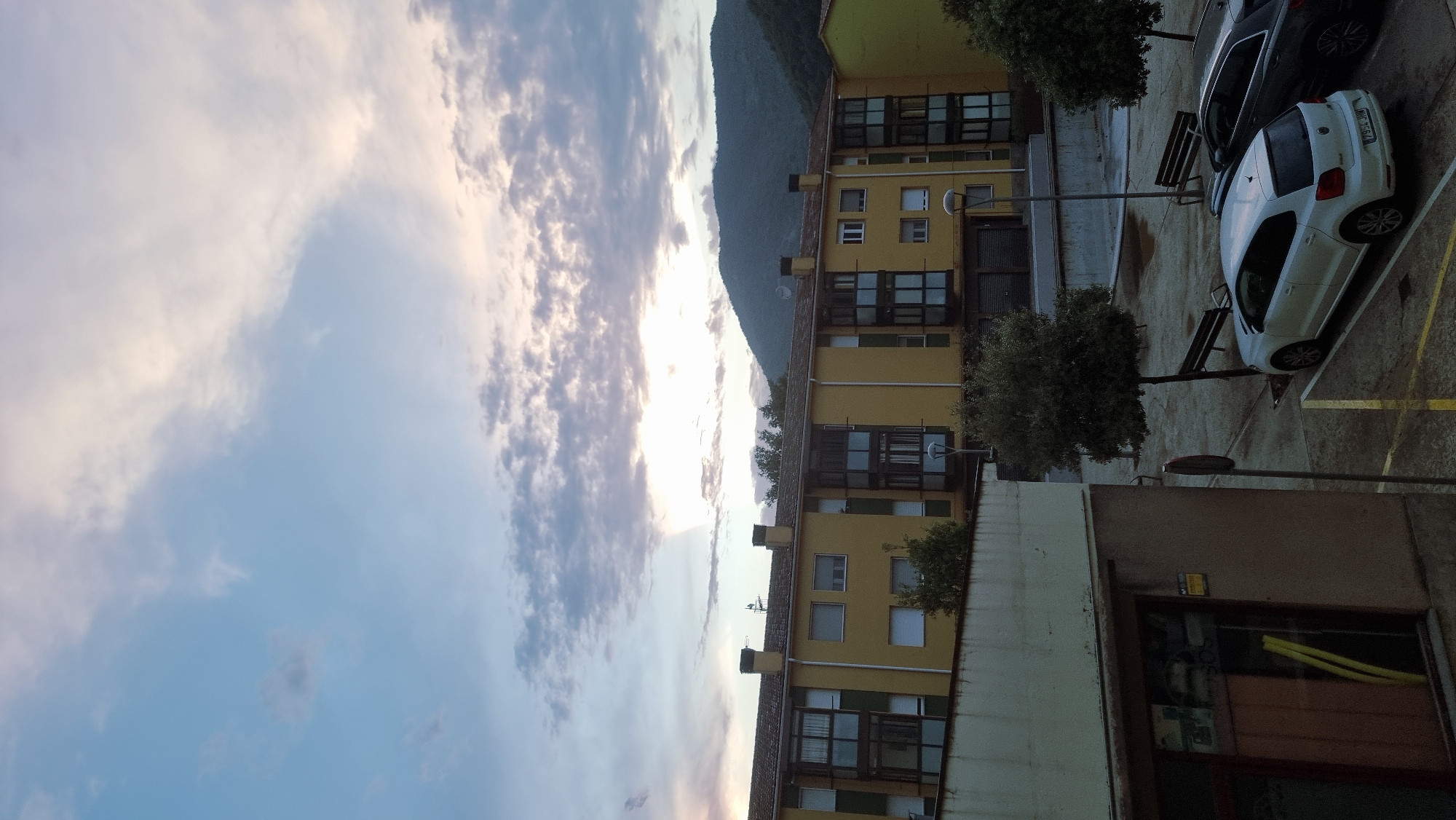 This is a full-frame, eye-level, medium shot of a building exterior in Sant Joan Les Fonts, Spain, captured during twilight. The sky is a mix of blue and purple clouds, with the sun peeking through from the upper center of the frame, illuminating the clouds. A large, yellow apartment building dominates the mid-ground, featuring multiple windows, balconies, and chimneys. In the foreground, to the left, is the side of a smaller building with a window displaying a reflective interior. The ground level includes a paved area with yellow parking lines, two parked cars (a white hatchback and a dark SUV), and several benches. Trees are scattered throughout the scene, one prominently in the mid-ground and two smaller ones on the right. A mountain range forms the background behind the apartment building. The overall impression is that of a quiet residential area at dusk.