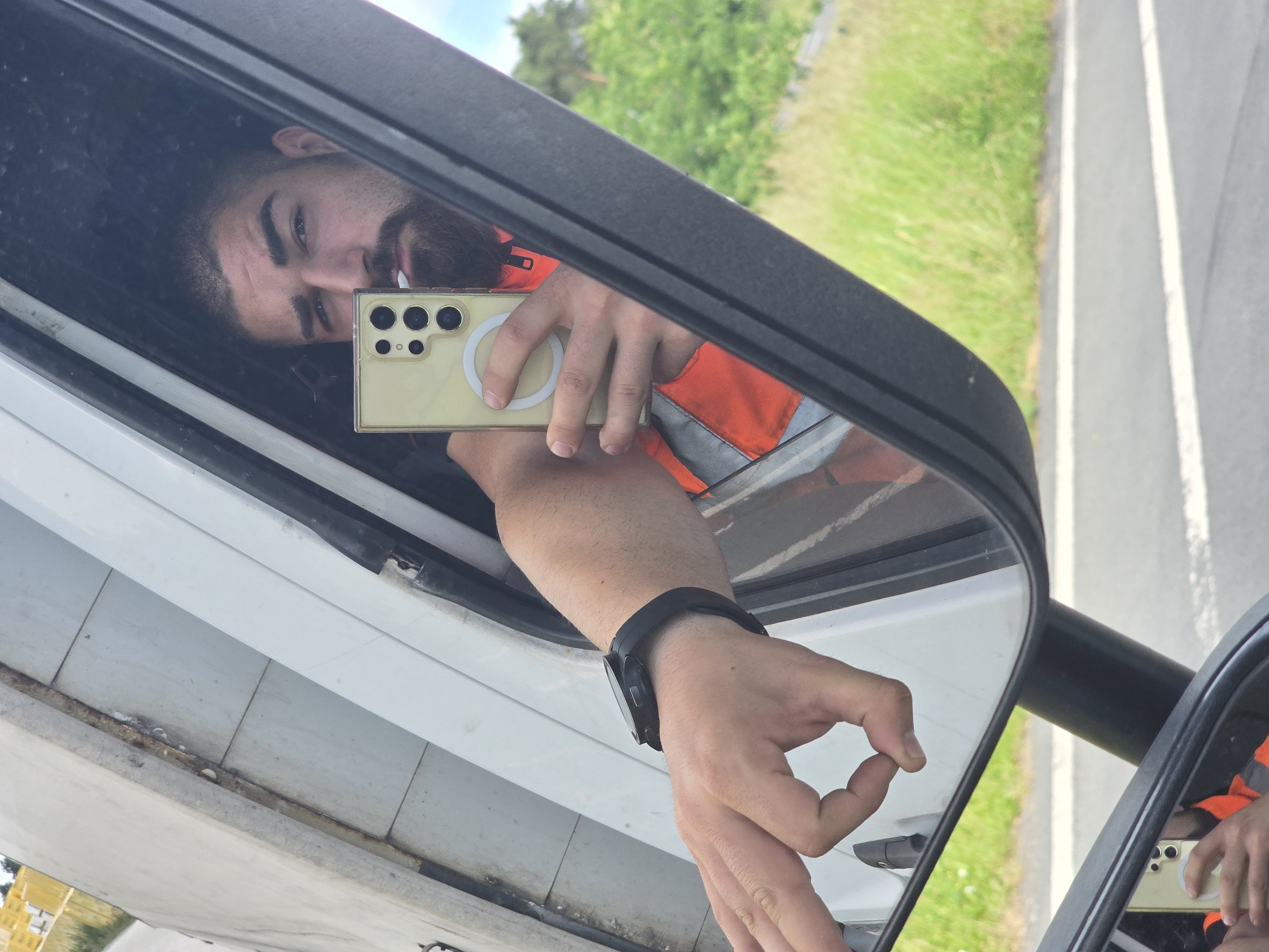 An eye-level selfie captures a man's reflection in a vehicle's side mirror, presenting a distorted and rotated perspective. The man, with a beard and dark hair, looks directly towards the camera of his smartphone, which he holds up with his right hand. The phone is yellow or gold, encased in a clear cover with a prominent circular MagSafe-like ring on the back, and multiple camera lenses are visible. He is wearing an orange reflective safety vest, indicative of outdoor or roadside work.
His left arm, adorned with a black smartwatch, extends out, and his hand forms an "OK" gesture. The vehicle's white exterior and part of its structure are visible around the mirror. The background, seen in the mirror's reflection, depicts a bright daytime scene of a road with a white line separating it from a stretch of green grass and distant trees under a clear sky. This suggests a location beside a rural or suburban road. The image is taken in Laußig, Germany. No discernible text is visible in the image.