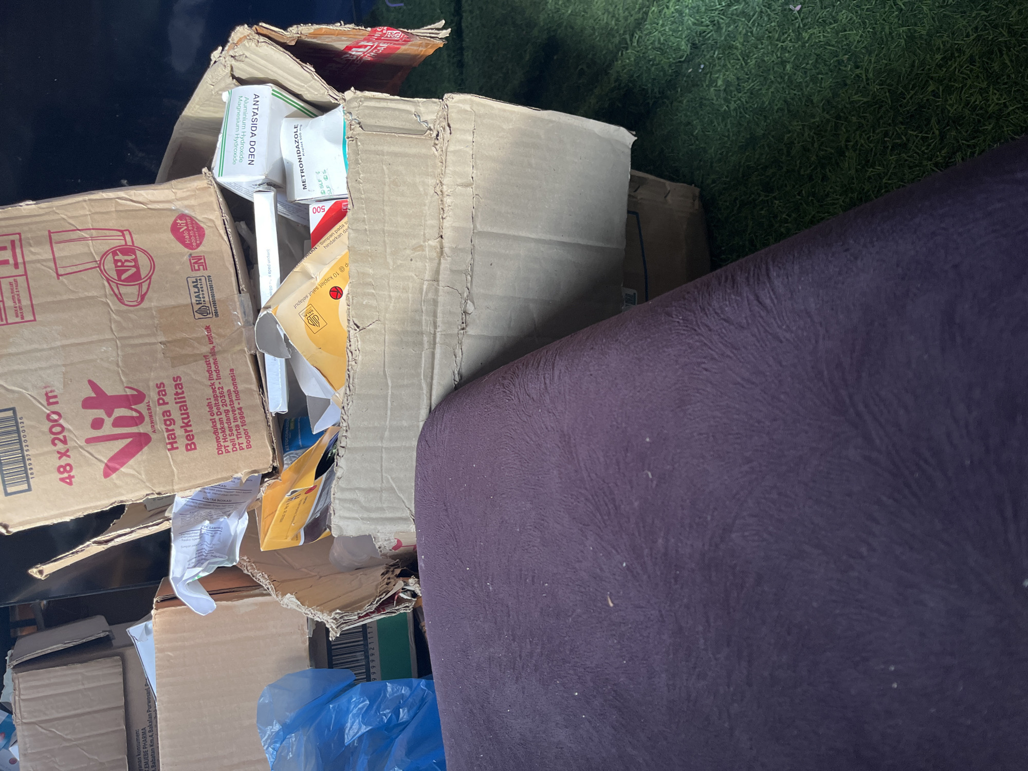 The image depicts a pile of cardboard boxes and packaging, likely in a storage or disposal area, with a portion of a dark purple upholstered object in the foreground and artificial green turf visible in the background. The boxes contain various items, including what appear to be pharmaceutical products.
One prominent box, branded "Vit AIR MINERAL," displays text in Indonesian stating "Harga Pas Berkualitas" (Fair Price Quality) and information about its production. Another box contains products labeled "ANTASIDA DOEN Magnesium Hydroxide" and "METRONIDAZOLE tablet 500 mg." Other visible packaging includes generic cardboard boxes, plastic bags, and yellow and orange paper packaging. The overall state of the items suggests they are either used, discarded, or awaiting organization.
The presence of Indonesian text on the packaging indicates the location is likely in Indonesia. While a specific address is not provided, if this scene were indeed in the city of Benuaraja, Indonesia, it would be a localized view within that municipality. The environment appears to be indoors or a covered outdoor space, given the lighting and the presence of artificial turf. There are no people or animals visible in the image.