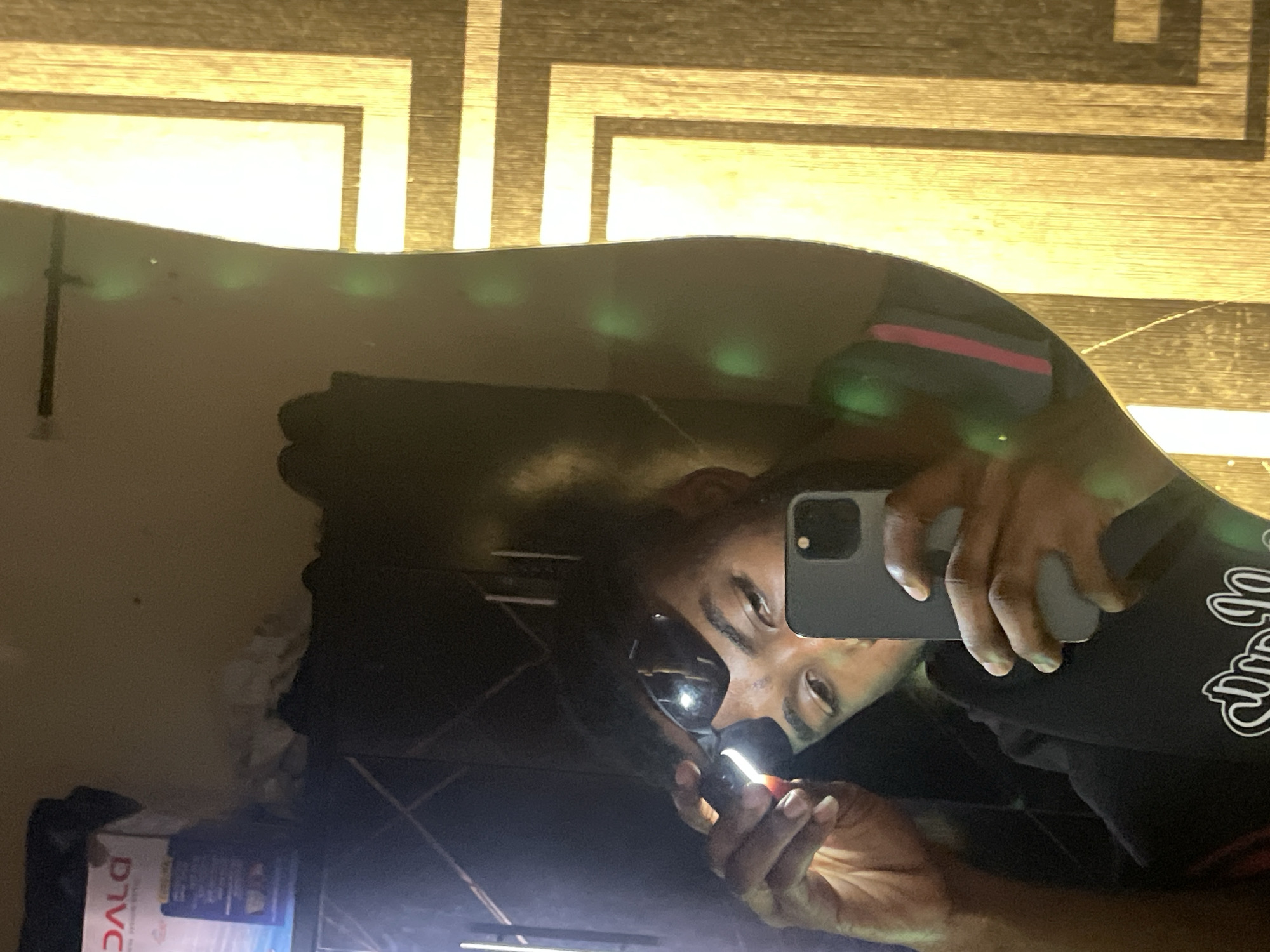 The image is a mirror selfie depicting one individual. The individual, appearing male with dark skin and dark hair, is reflected in an irregularly shaped mirror, holding a gray smartphone with a prominent camera bump in his right hand to capture the image. His left hand is lifting a pair of dark-lensed sunglasses from his face, partially revealing his eyes and eyebrows. He is wearing a dark garment, possibly a t-shirt or hoodie, with a white graphic design visible on the lower left side.
In the upper left portion of the mirror's reflection, an inverted white and blue box, partially labeled "DALO," rests on a dark surface, likely a shelf or cabinet. Behind this, a light-brown wall shows obscured, possibly rumpled, objects. The reflected background is generally dim.
To the right of the mirror, a wall or surface with a distinct geometric pattern of interlocking vertical and horizontal lines, resembling wood grain or a patterned panel, is brightly illuminated by a warm, yellow light. Multiple green, circular lens flares are visible across the mirror surface and the adjacent wall, suggesting interaction with a light source. No visual elements within the image confirm the location as Lokoja, Nigeria.