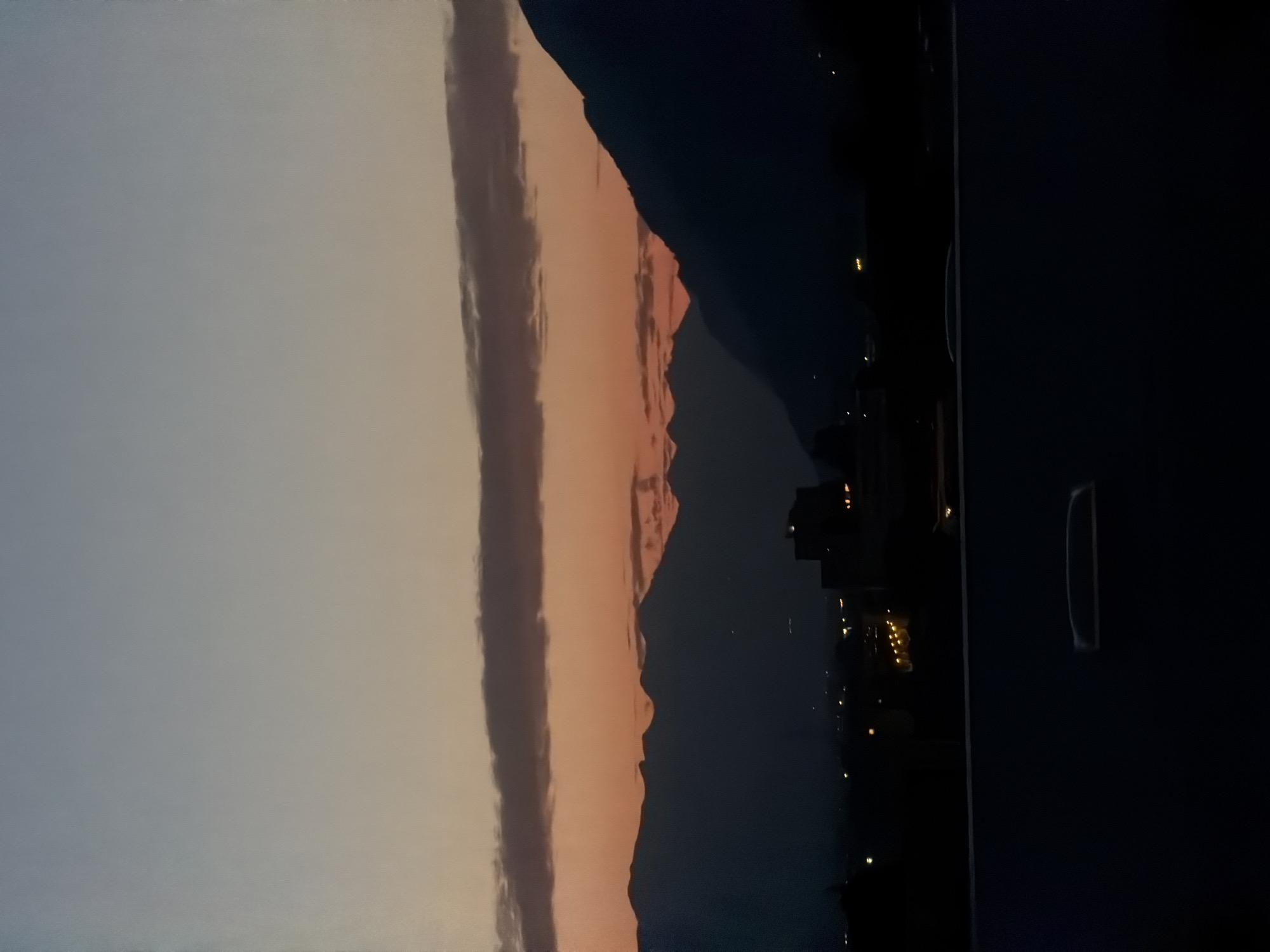 This image captures a dusk scene with a silhouetted mountain range against a gradient sky, transitioning from pale blue at the top to orange near the horizon. A horizontal band of clouds is visible across the middle of the sky. In the foreground, the dark silhouettes of buildings and lights suggest an urban area, likely Martigny, Switzerland, nestled at the base of the mountains. The bottom portion of the image is dominated by a dark, flat surface, possibly a road or balcony, with a reflective element visible on it.