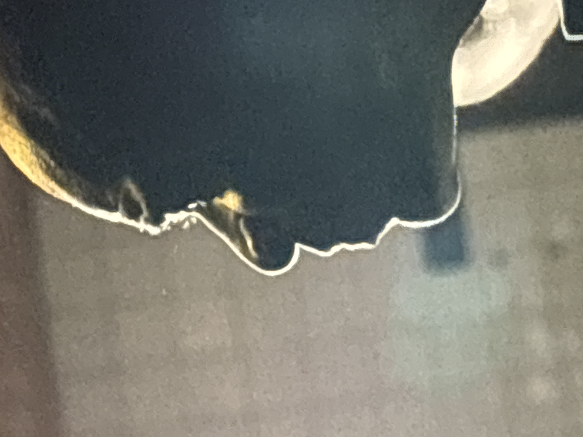 This is a close-up shot of the underside of an object, likely metal or plastic, which has a dark color and a lighter-colored, possibly chrome, trim along the edge. A drip or protrusion is visible along the lower edge. The background is a blurry, neutral tone. There is no information within the image to confirm the location in Budva, Montenegro.