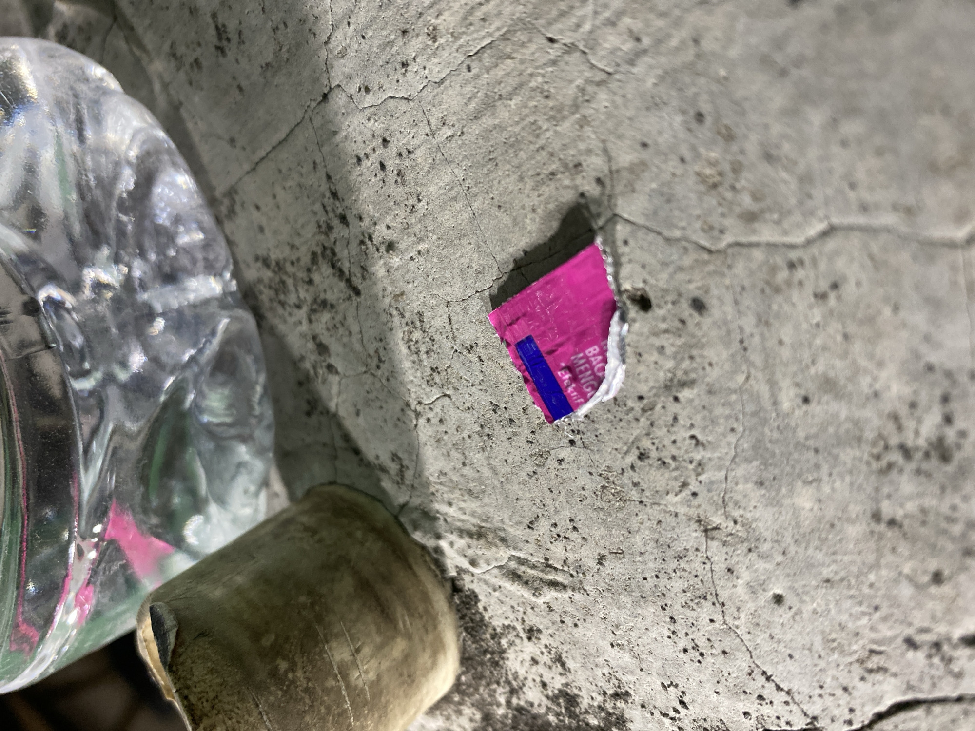 The image depicts a close-up, high-angle shot of a small, torn piece of pink and blue wrapper on a cracked concrete surface. The wrapper appears to be from a food item or candy, with some text partially visible. In the background, out of focus, is a glass container filled with a clear liquid, possibly water, and what appears to be a beige, cylindrical object, possibly a cup or a piece of pottery. The lighting suggests an outdoor setting, possibly at night due to the harsh shadows and focus. The specific location in Kasihan, Indonesia, is not discernible from the image itself, as there are no identifying landmarks or contextual clues.