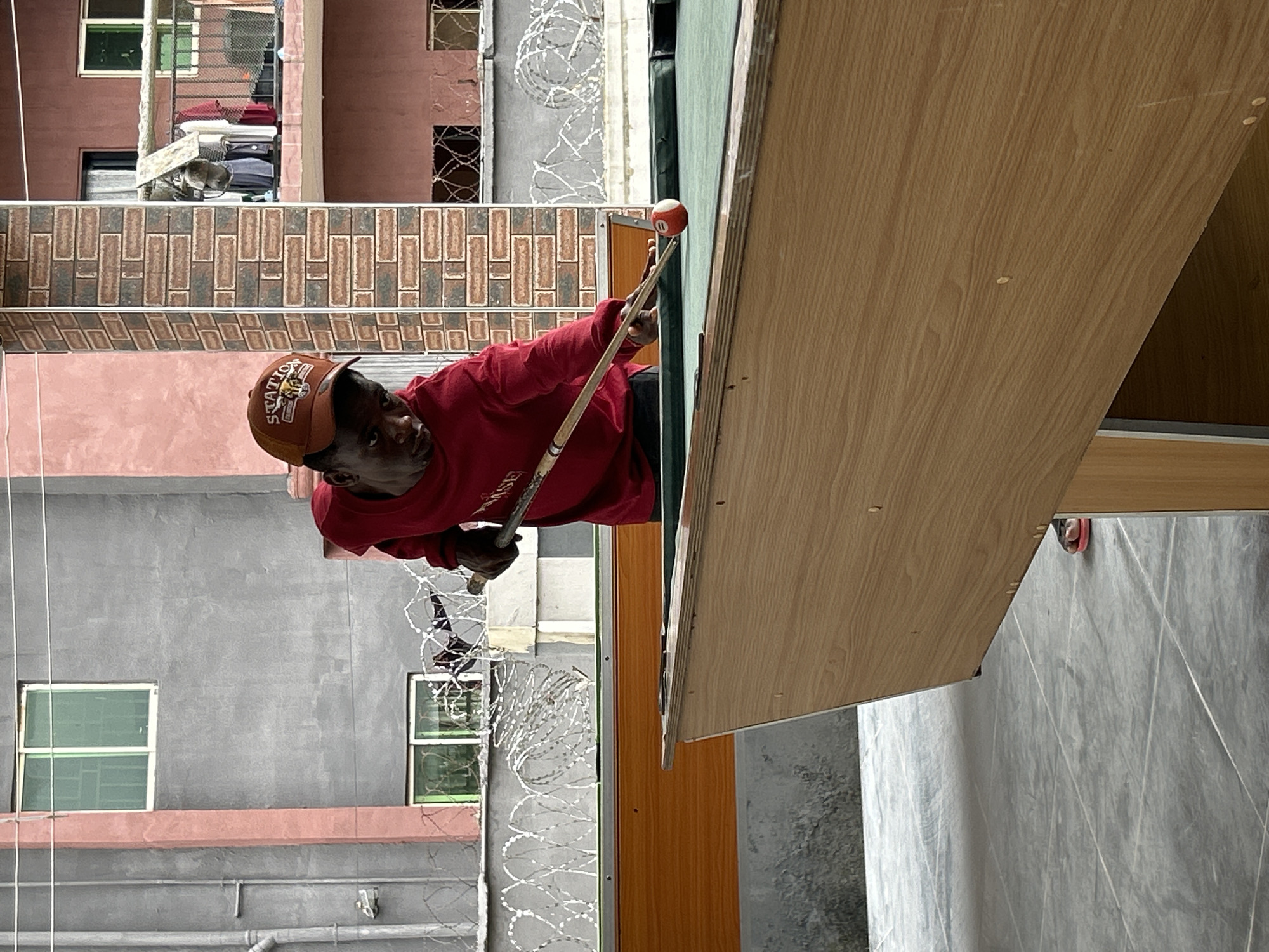 The image shows a young Black male playing billiards outdoors in Ihiagwa, Nigeria. He is wearing a red long-sleeved shirt and a brown baseball cap with "STATION" written on it. He is focused on hitting a red billiard ball with a cue stick. The billiard table has a green felt surface and a wooden frame. In the background, there is a grey building with pink trim, a brick building, and a balcony with laundry hanging. Razor wire fencing is visible along a wall. The ground is paved with grey tiles. A pair of red flip-flops are visible near the billiard table.