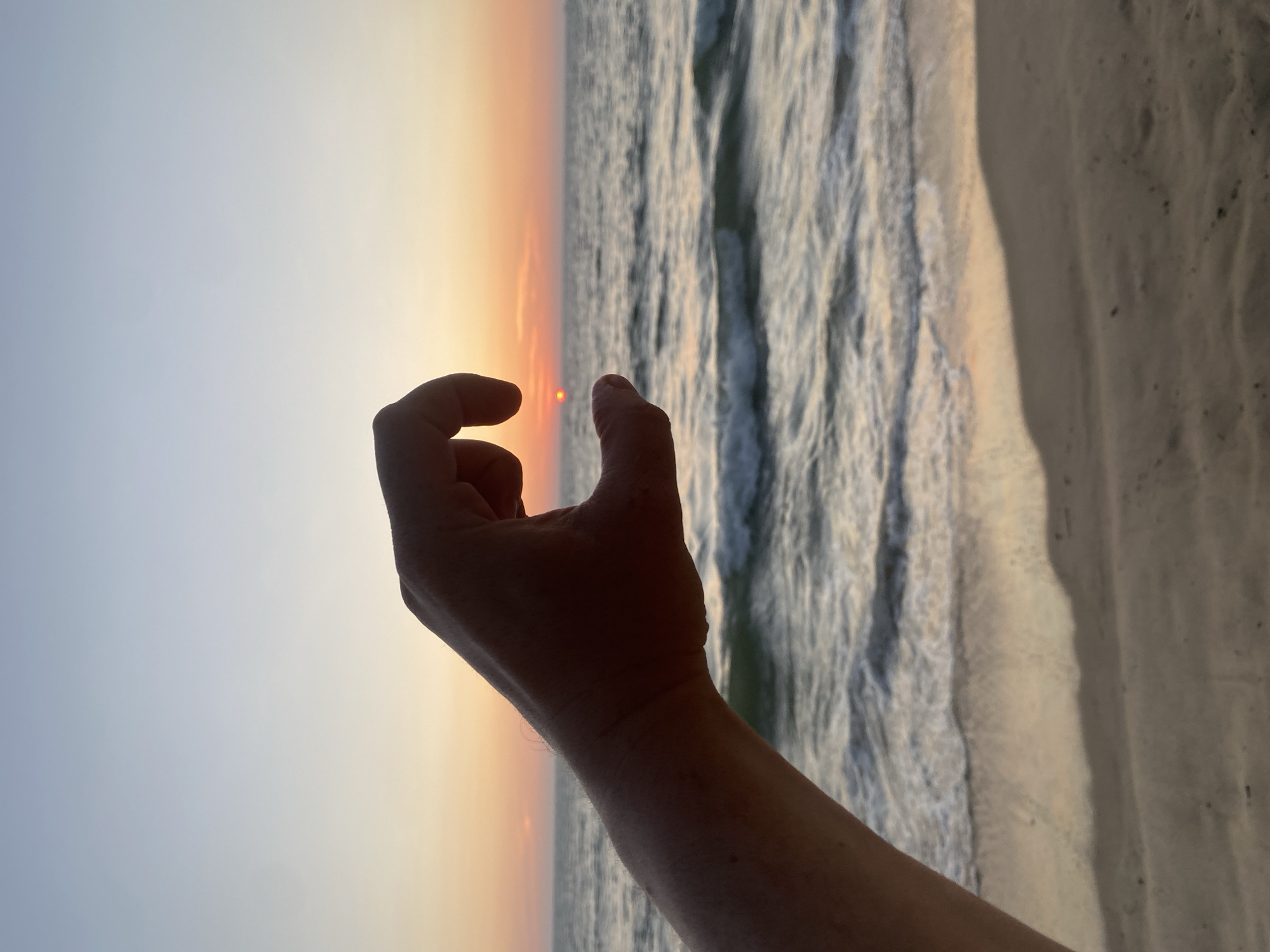 A person's arm and hand reach toward the sunset over the ocean. The hand is curved with the thumb and pointer finger close together, appearing to "hold" the sun. The sky transitions from blue to orange as the sun sets. The ocean waves reflect the light. The scene is likely a beach, with a sandy shoreline visible on the right side of the frame. The photo provides no evidence of the image being taken in Choeng Thale, Thailand.